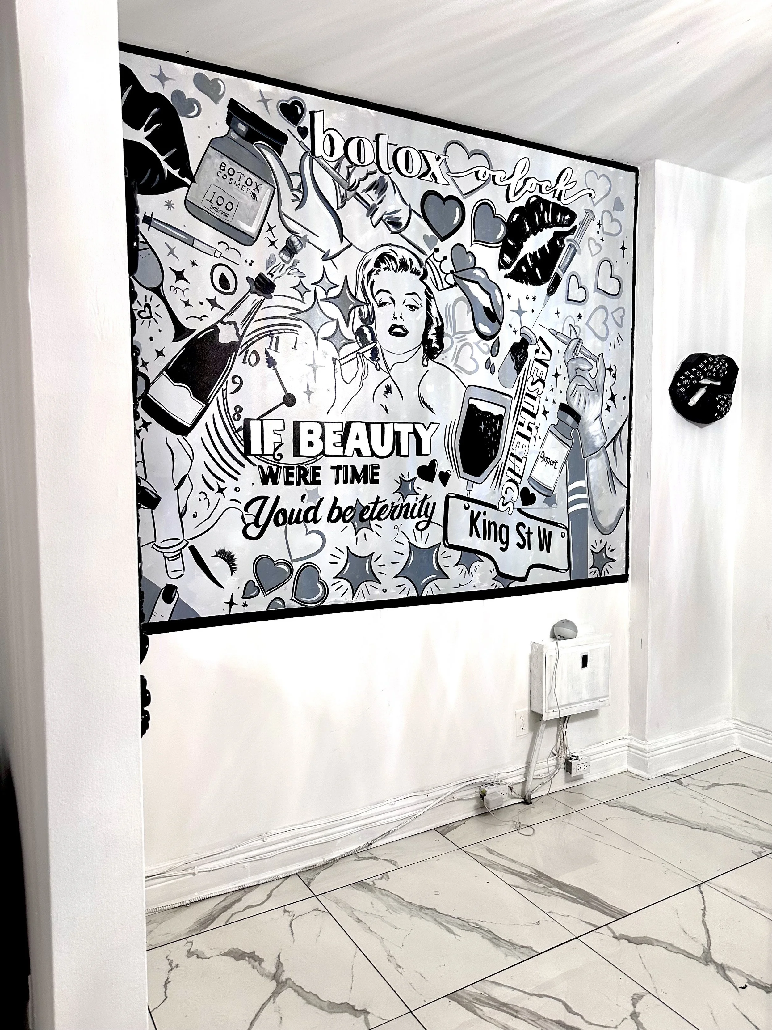 A black and white mural with vintage beauty and cosmetic-related illustrations and sayings, including a woman, hearts, lips, bottles, and the phrase 'beauty was time you'd be eternity,' on a white wall in a room with tile flooring.
