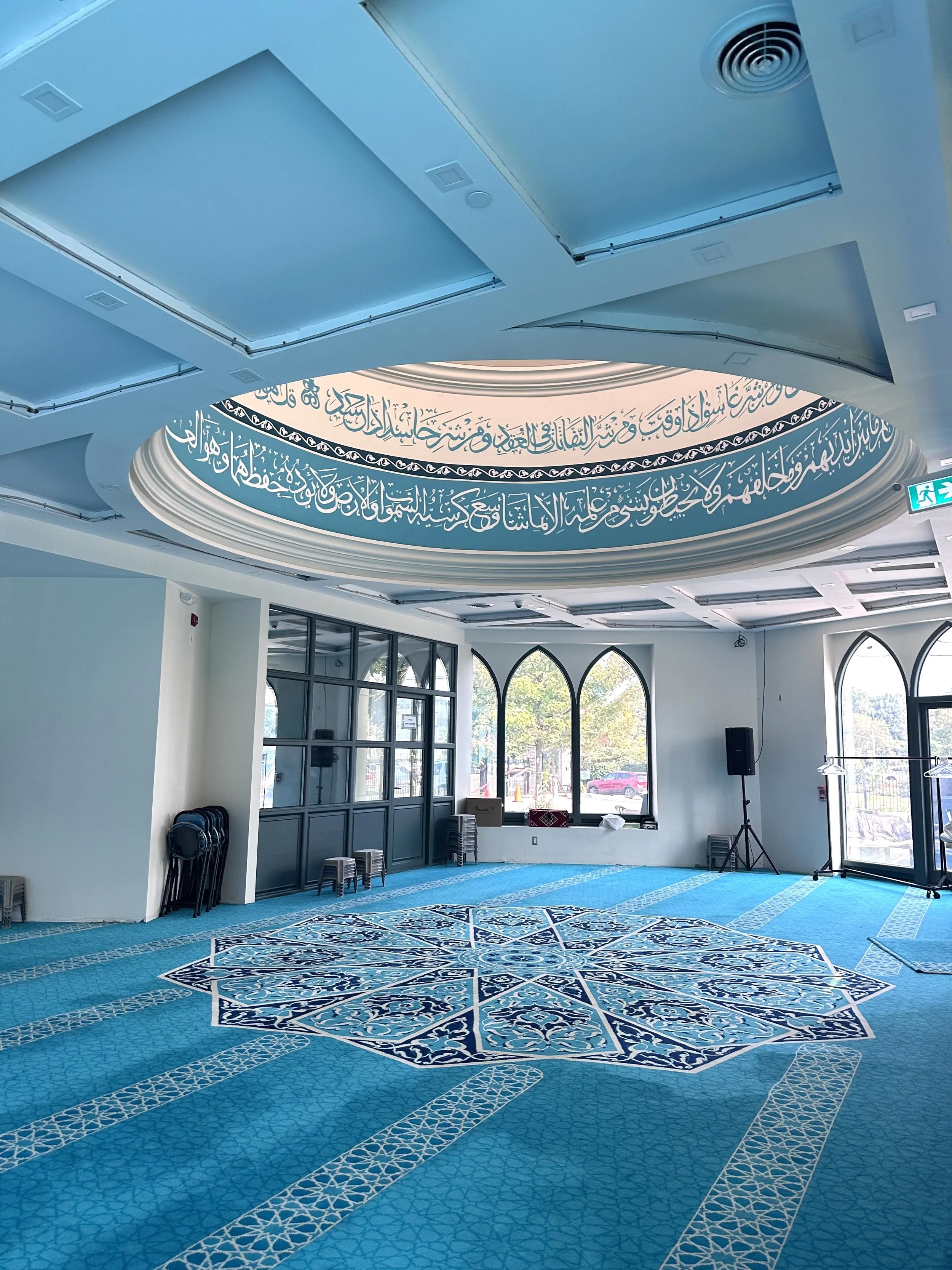 Interior of a prayer room with blue carpet, stained glass windows, and Arabic calligraphy on a circular ceiling design.