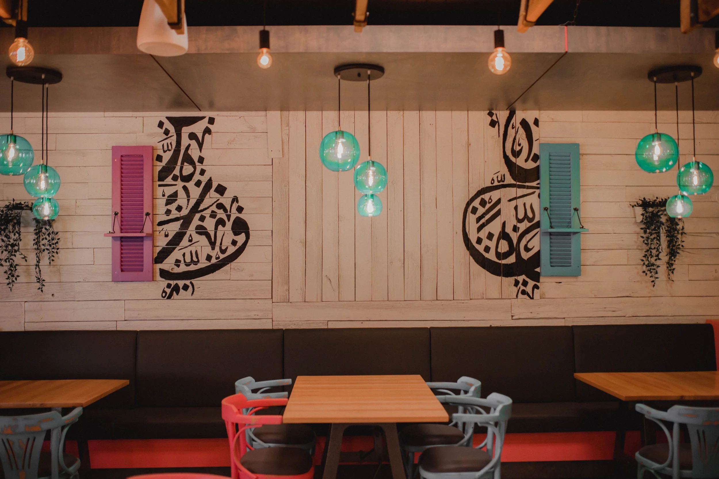 Interior of a modern restaurant with wooden walls, colorful shutters, hanging globe lights, and Arabic calligraphy art.