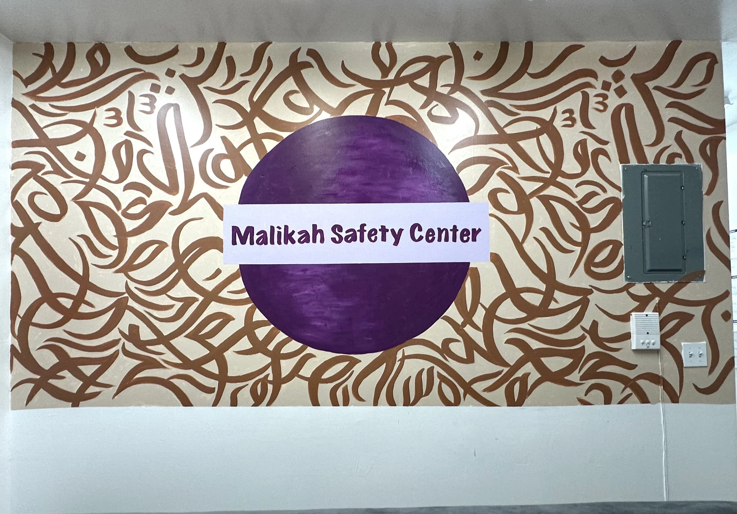 Wall with a patterned background, purple circle in the center with a white banner reading 'Malikah Safety Center'.