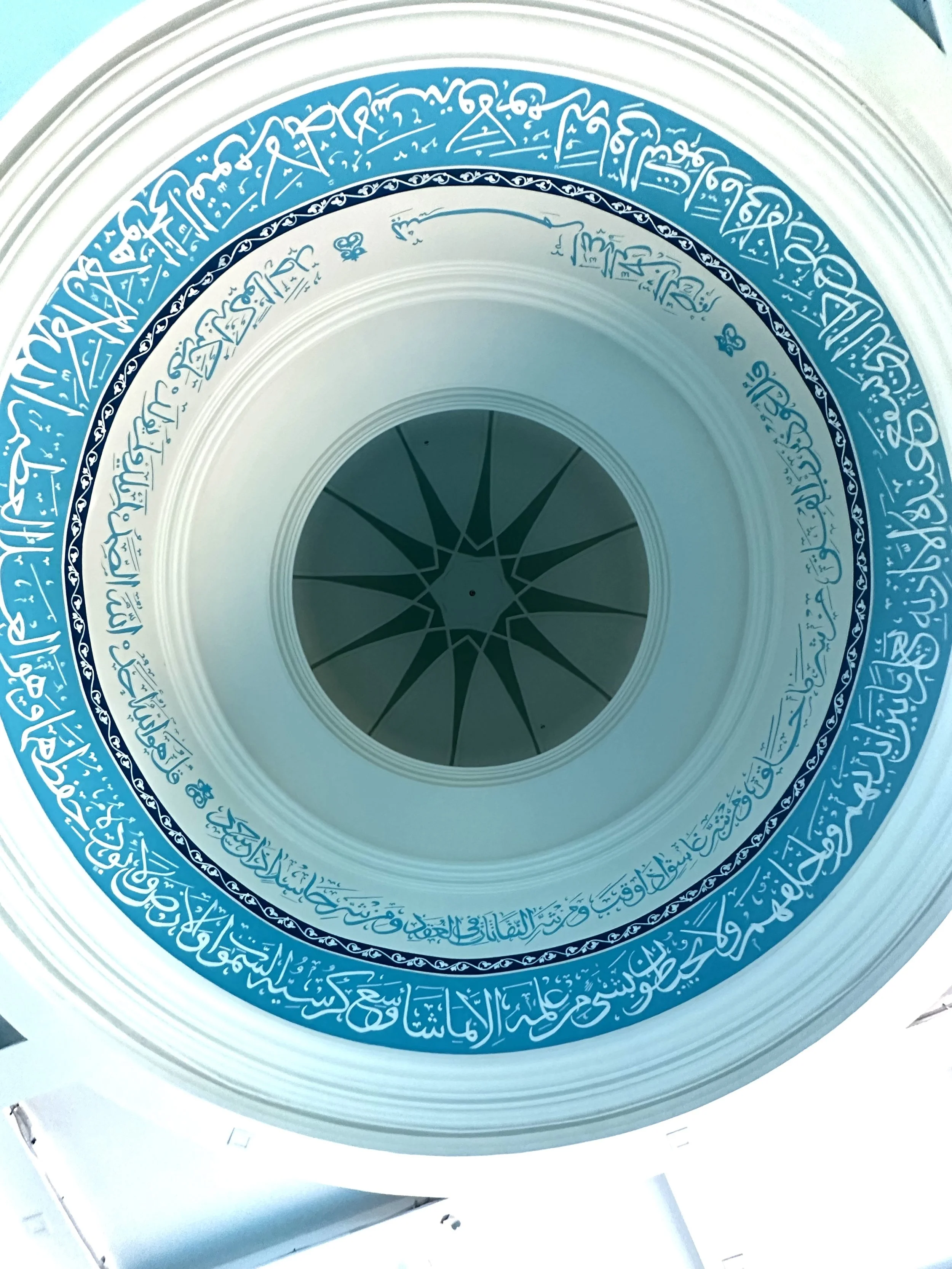 Inside view of a dome ceiling with intricate blue and white Islamic calligraphy around the perimeter and a geometric star pattern in the center.