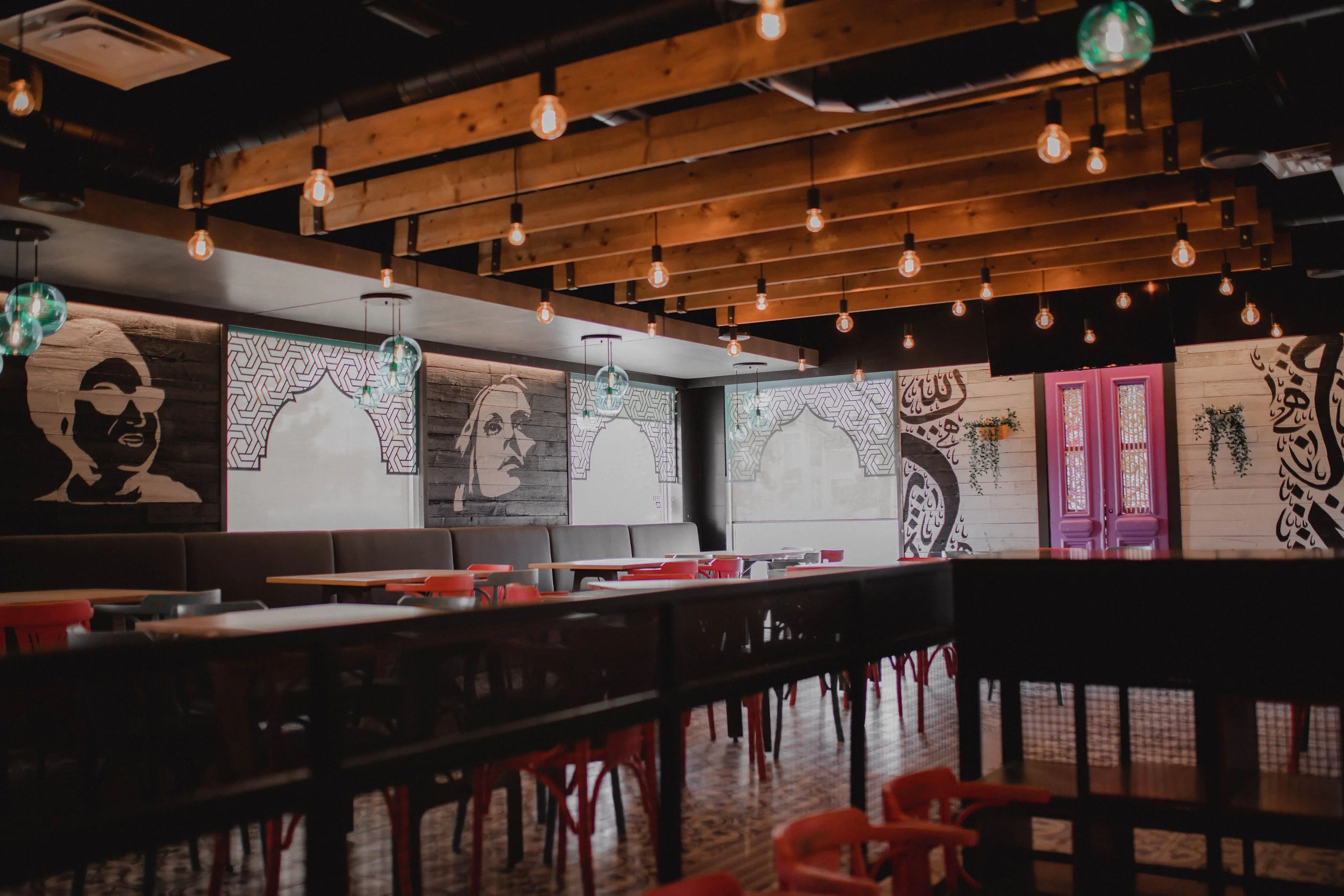Empty restaurant with wooden ceiling beams, hanging Edison-style light bulbs, and wall art featuring portraits and Arabic calligraphy.