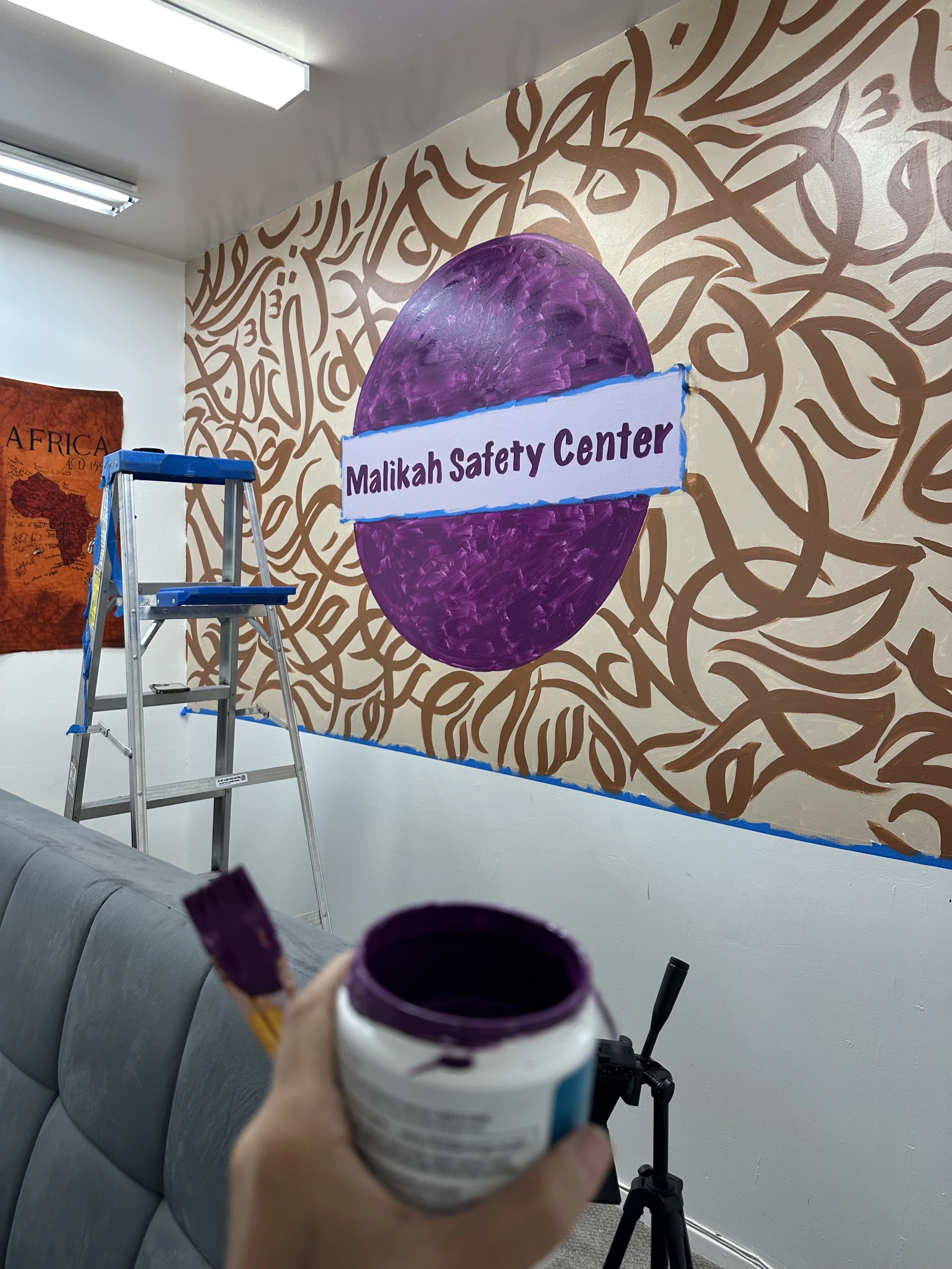 A person holding purple paint and a brush, painting a wall with a large purple circle and the words 'Malikah Safety Center' in purple on a white banner. The wall has a decorative brown pattern background. An aluminum ladder is standing upright nearby