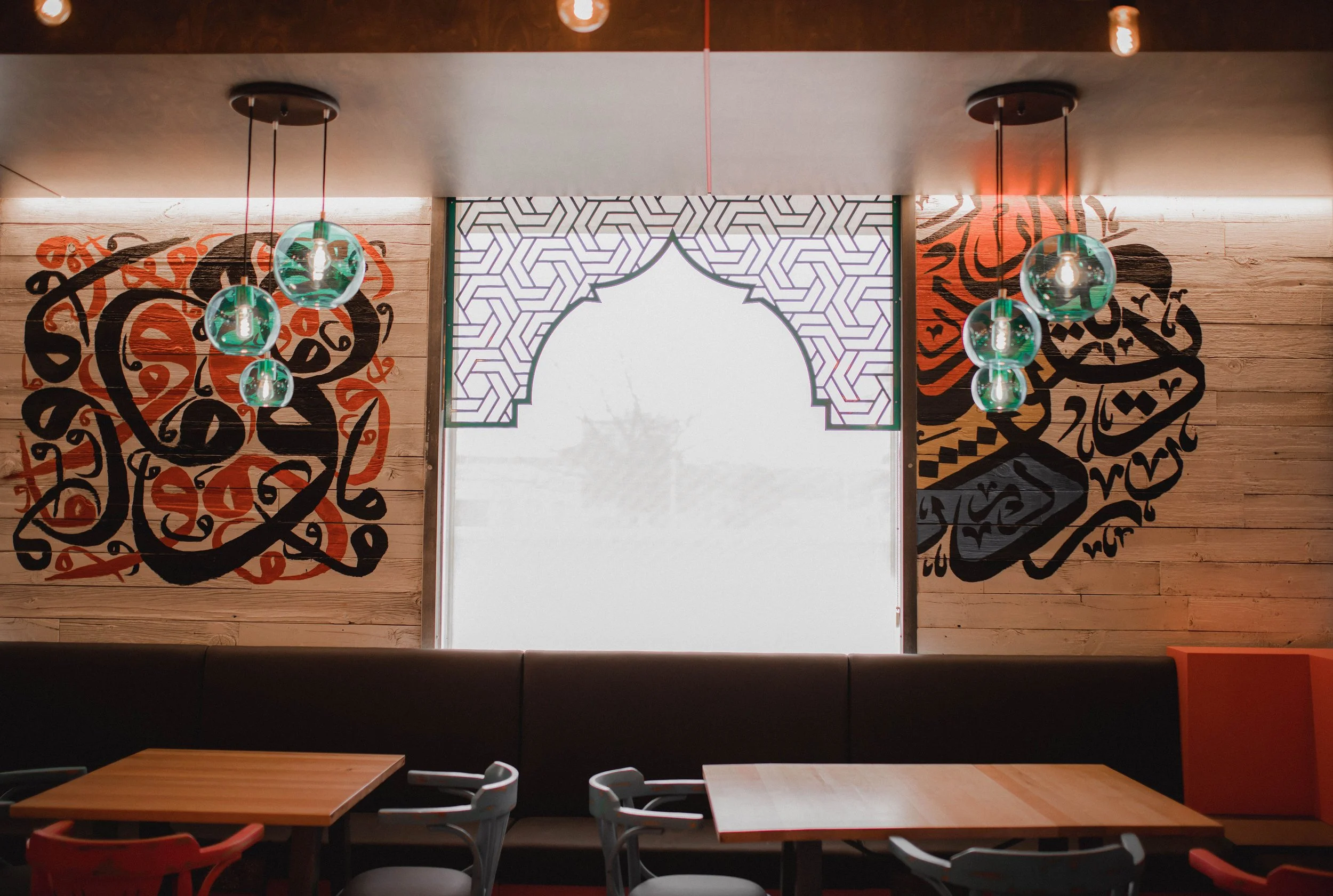 Interior of a restaurant with wooden wall panels, decorative black and orange calligraphy art, modern pendant lights with glass globes, and empty tables and chairs.