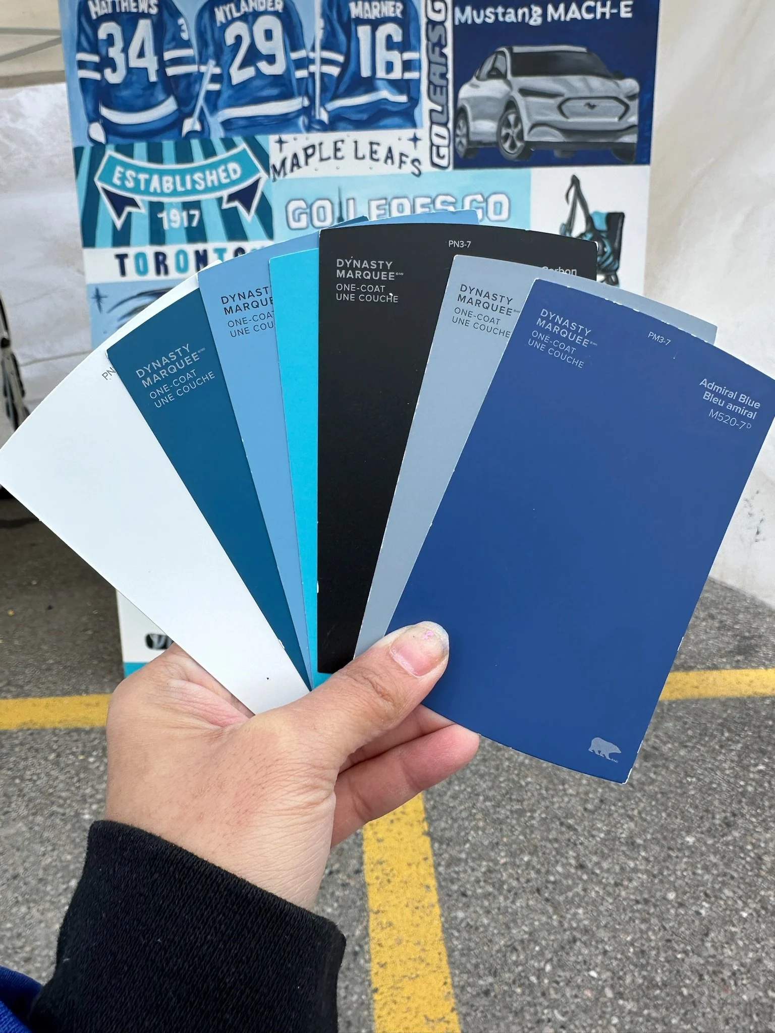 A hand holding a fan of paint color swatches, with a Toronto Maple Leafs poster in the background.