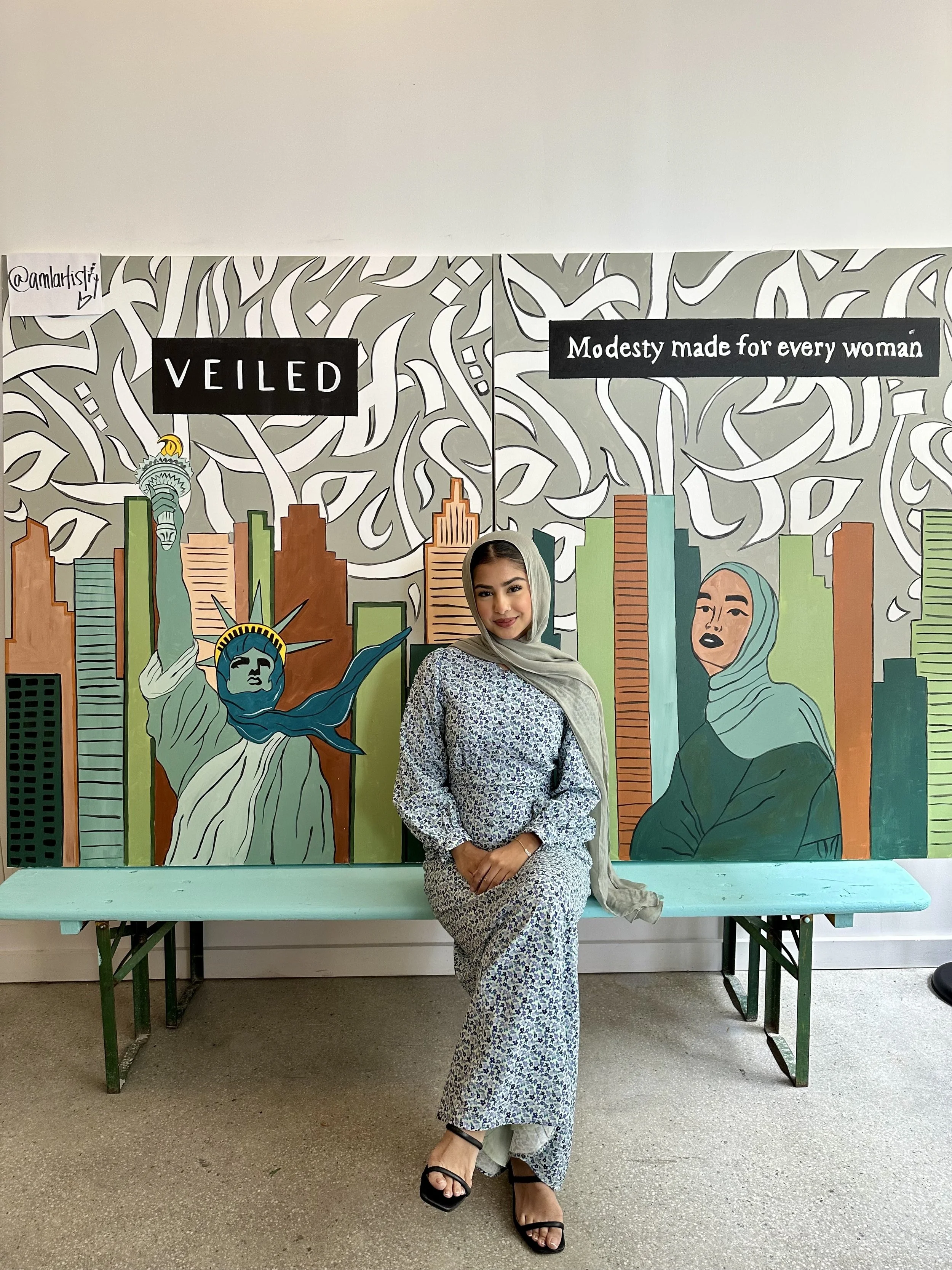 A woman wearing a patterned dress and heels sitting on a bench in front of a mural featuring the Statue of Liberty and a woman in a hijab with city buildings in the background. The mural has text reading 'Veiled' and 'Modesty made for every woman'.