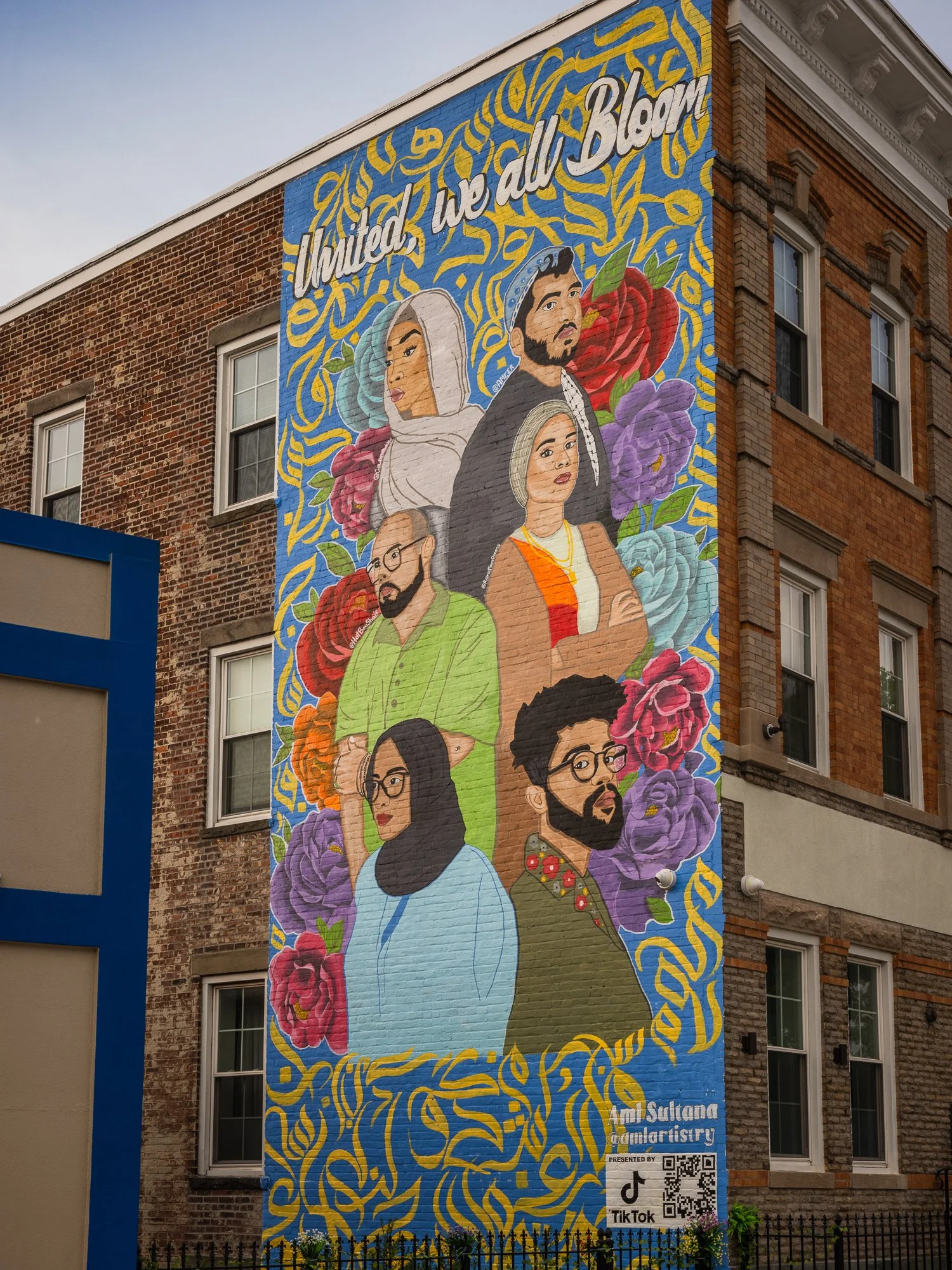 Colorful mural of diverse people with flowers and the phrase 'United we all bloom' on a brick building wall.