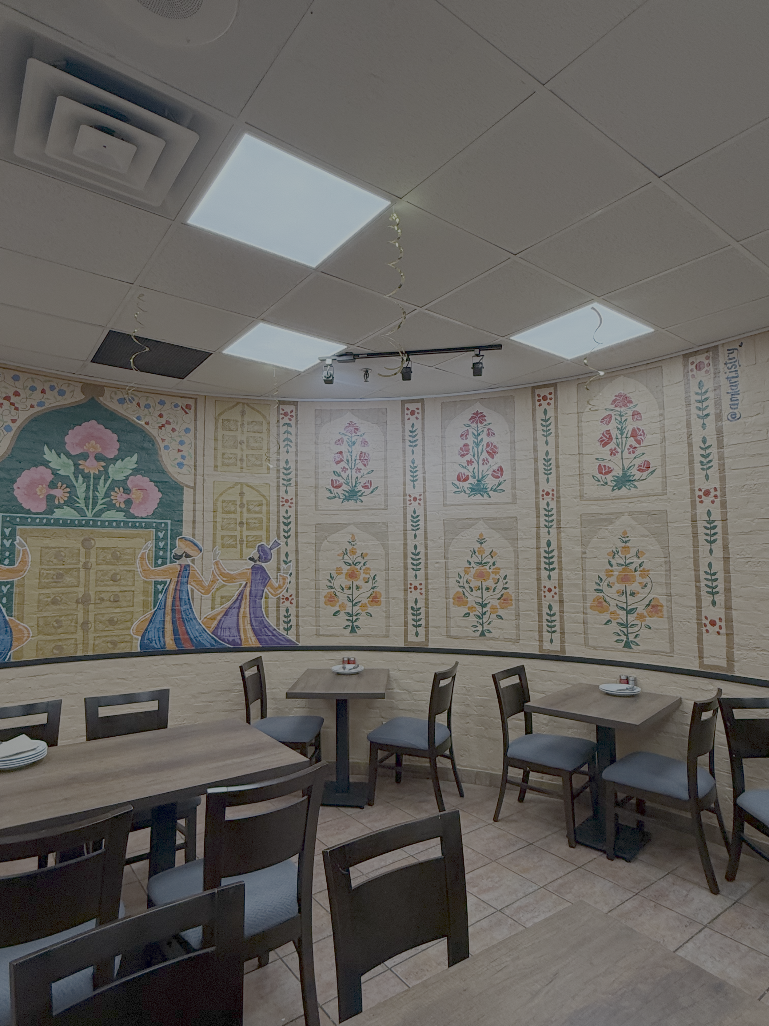 Empty dining area with wooden tables and chairs, and a decorative mural with floral and dancing figures on the wall.