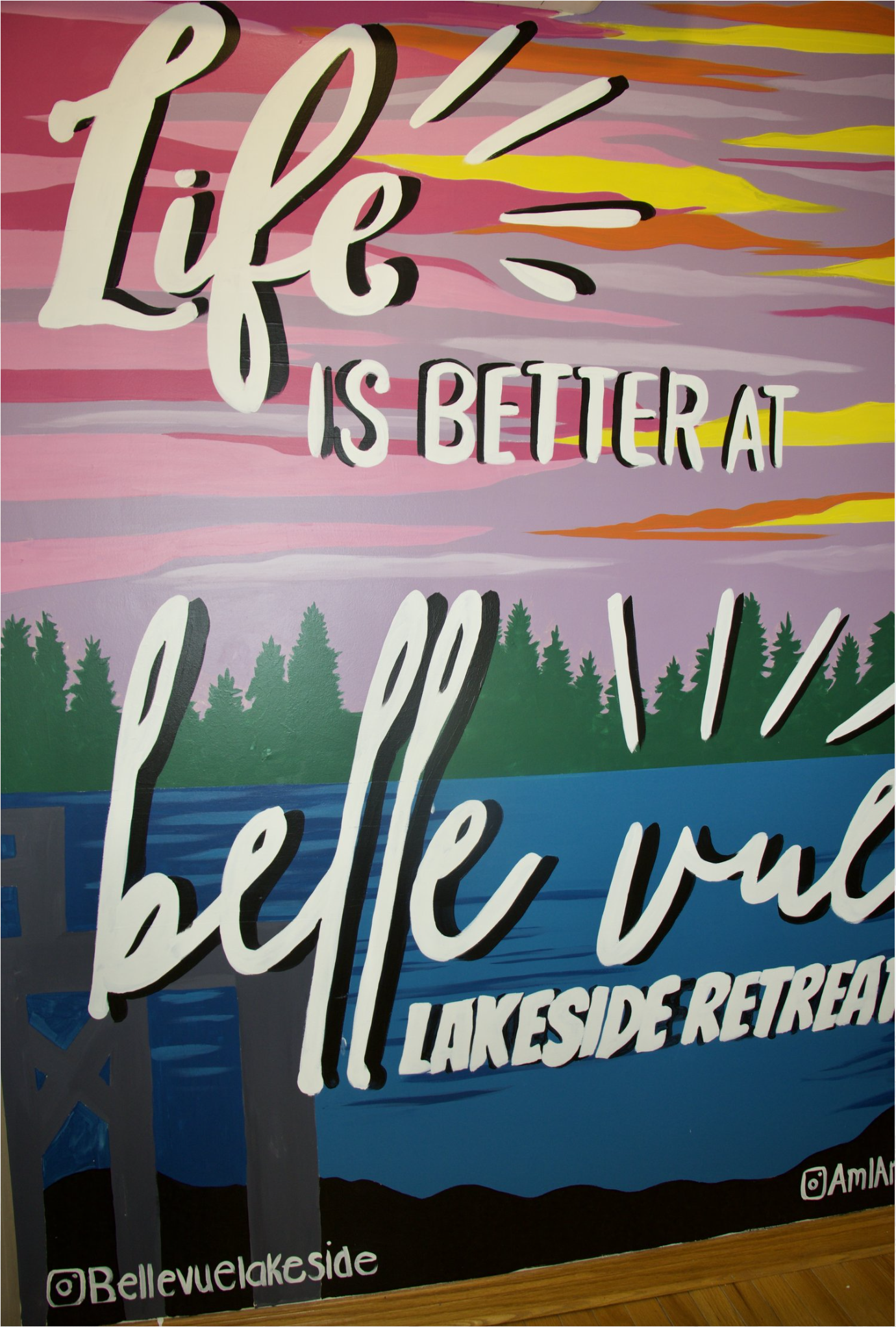 Colorful mural with sunset sky, forest, and lake, featuring the text 'Life is better at Bellevue Lakeside Retreat' in large, artistic lettering.