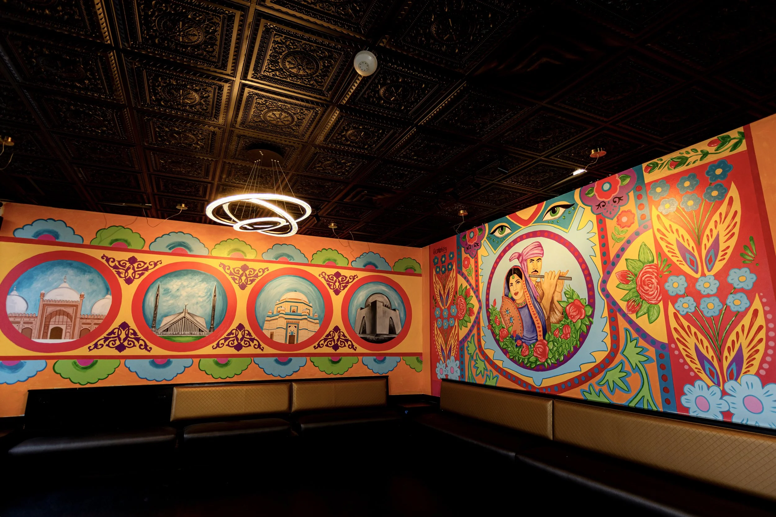 Colorful Indian-themed mural inside a room with dark ornate ceiling and modern circular chandelier, featuring traditional architecture, floral patterns, and a woman playing a flute.