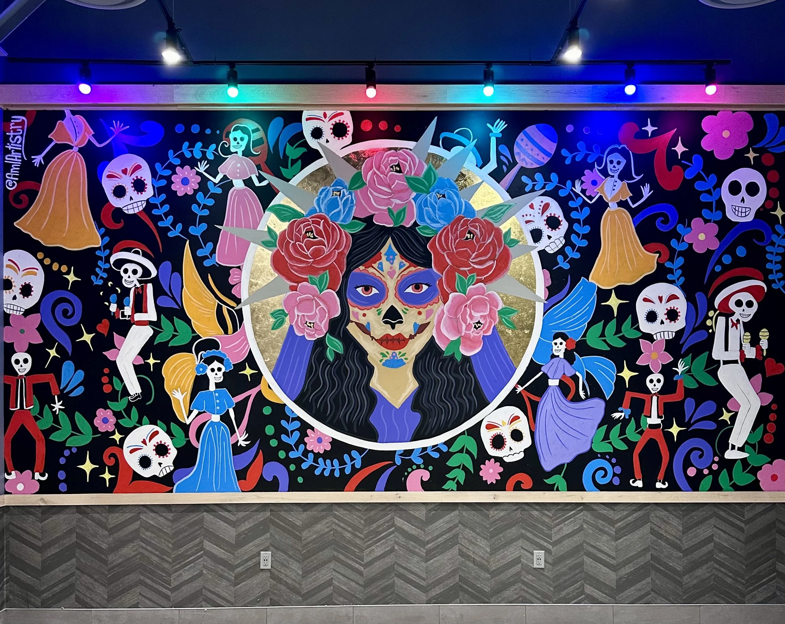 Colorful mural featuring a central woman with a skull face, flower crown, and painted face, surrounded by skeleton figures, flowers, and festive elements in bright colors against a dark background.