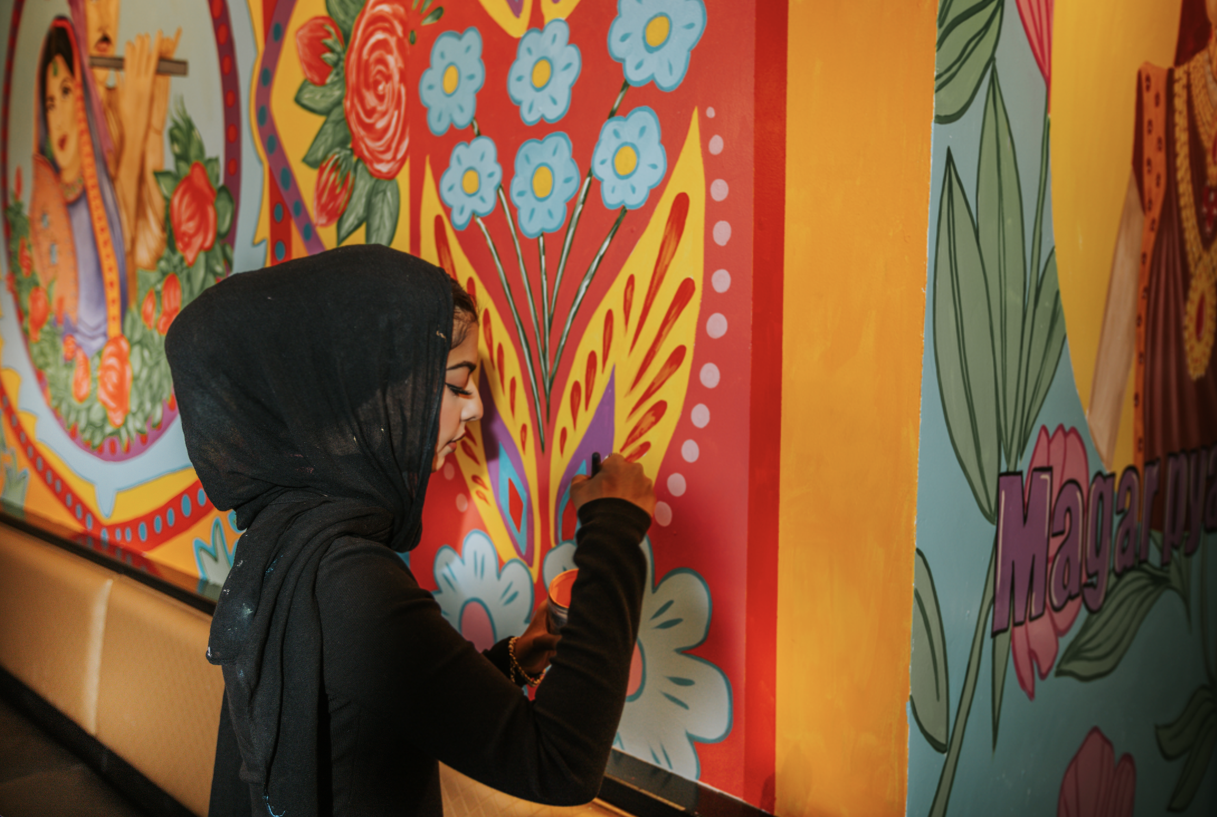 A woman wearing a black hoodie and a black headscarf is painting a large colorful mural with vibrant flowers and patterns.