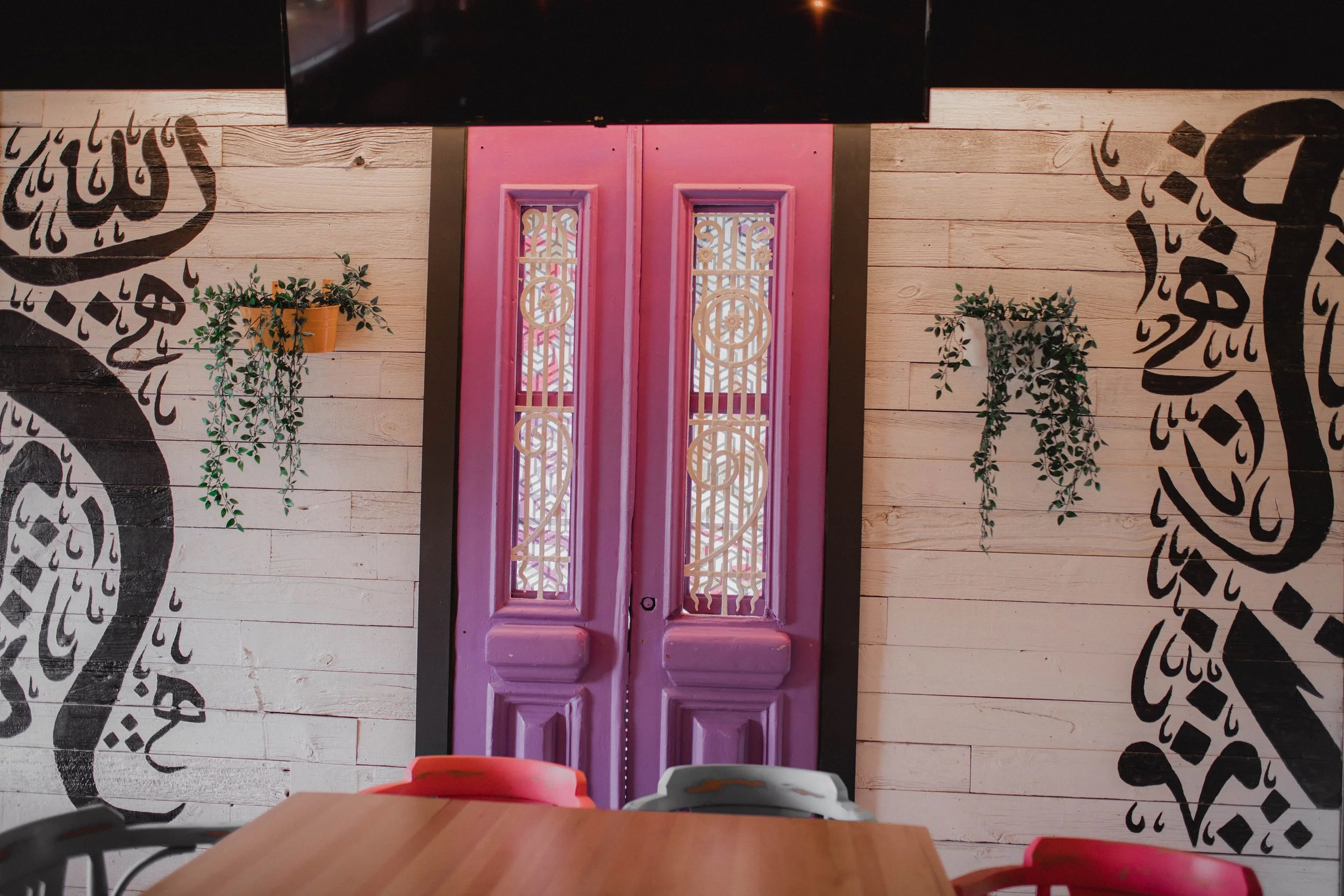 Pink double doors with intricate metal grilles, flanked by white wooden walls with black Arabic calligraphy and hanging green plants, in a café or restaurant setting.
