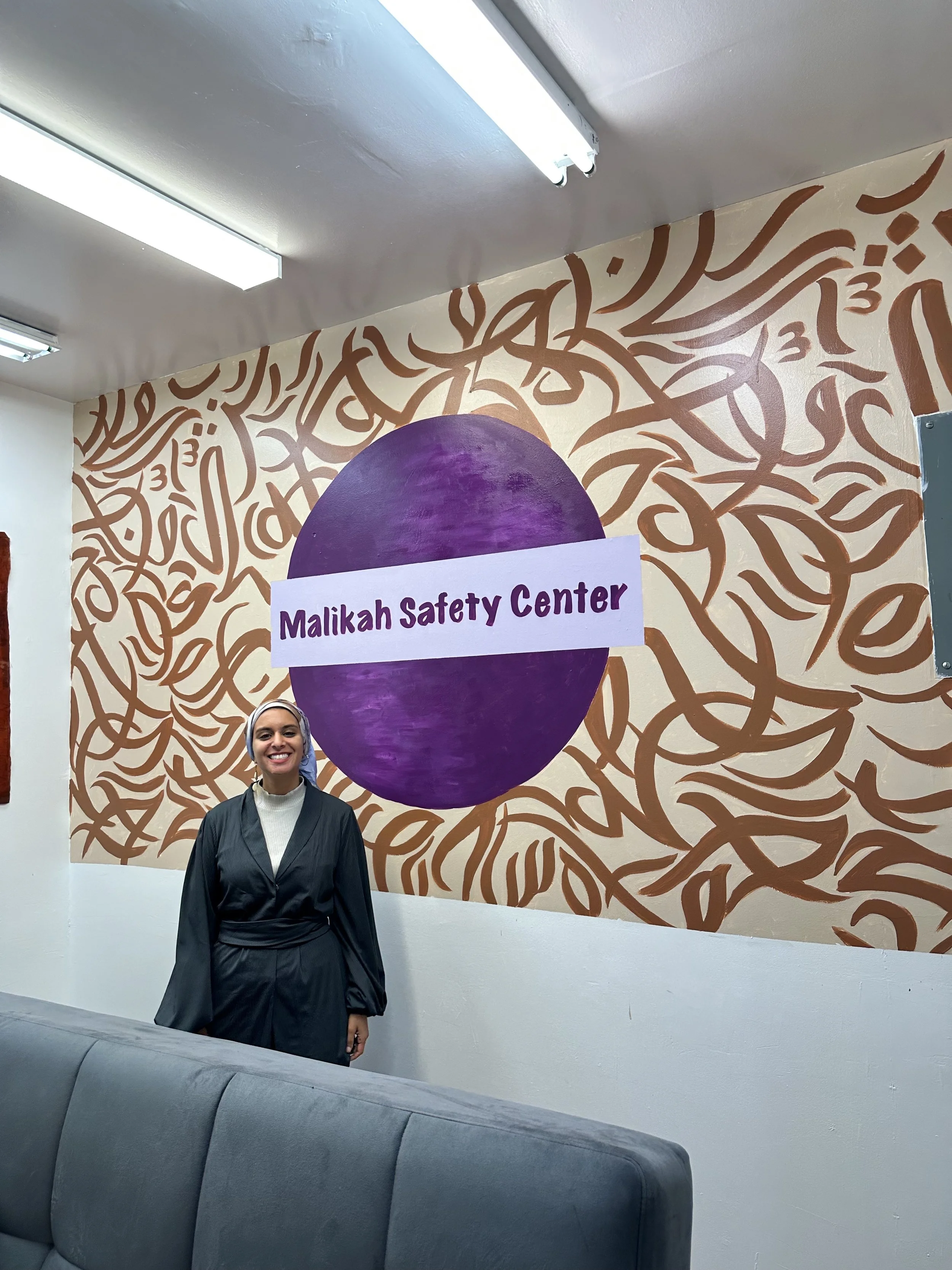 A woman wearing a hijab and black clothing, smiling, standing in front of a mural with a purple circle and the words 'Malikah Safety Center' on it.