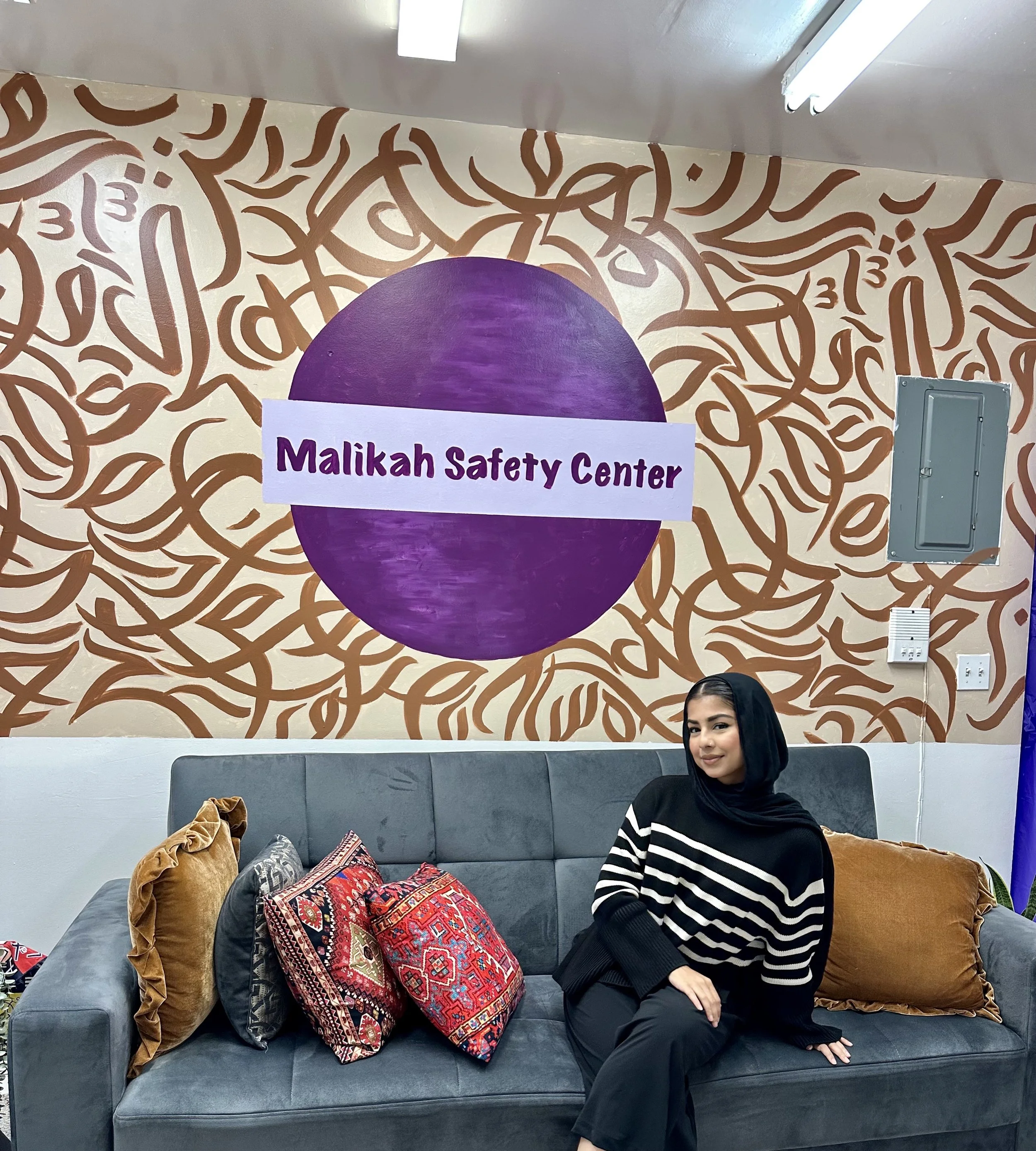 A woman sitting on a gray sofa with assorted colorful pillows, in front of a wall with a large purple circle painted on it and the words "Malikah Safety Center" written across the circle. The woman is wearing a black and white striped sweater and a b