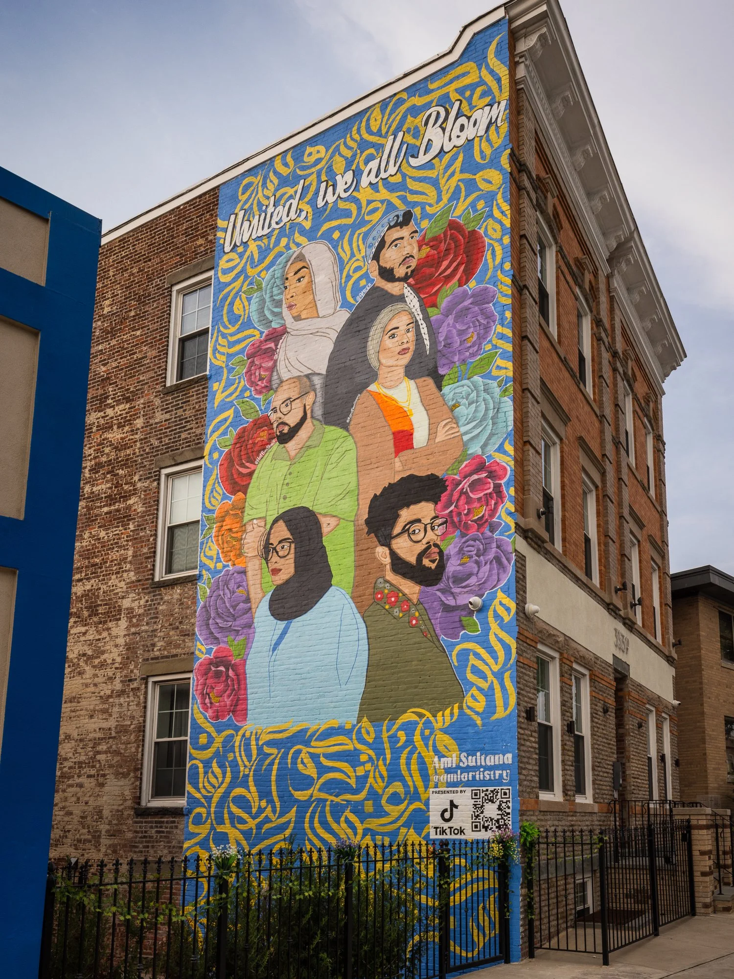Colorful mural on the side of a building depicting diverse portraits of people surrounded by flowers with the phrase "United, We All Bloom" at the top, and a QR code and TikTok logo at the bottom.