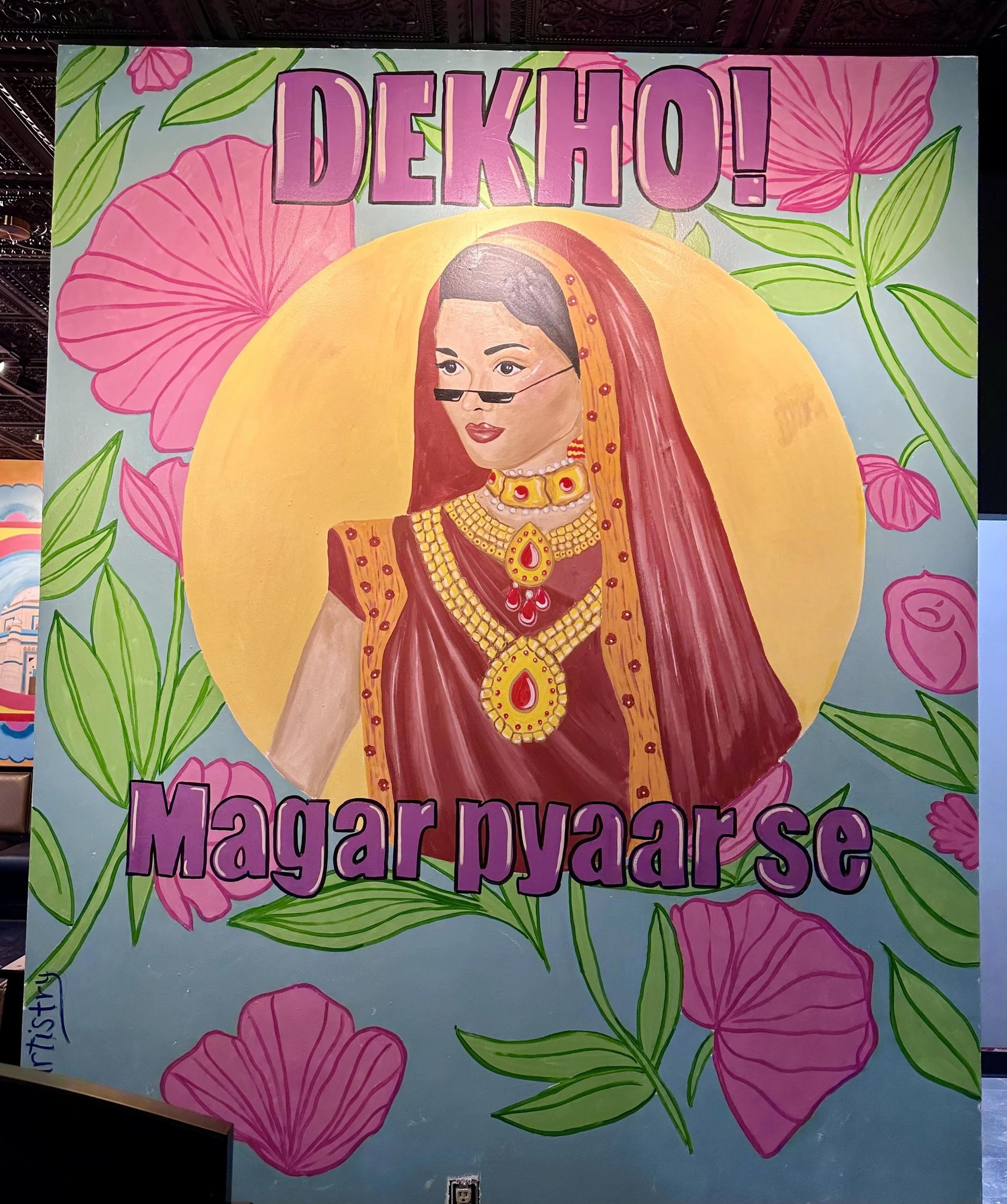 Colorful mural illustration of a woman in traditional Indian attire, adorned with jewelry, glasses, and a red and yellow saree, against a background of pink flowers and green leaves, with the Hindi words 'DEKHO! Magar pyaar se' in purple text at the
