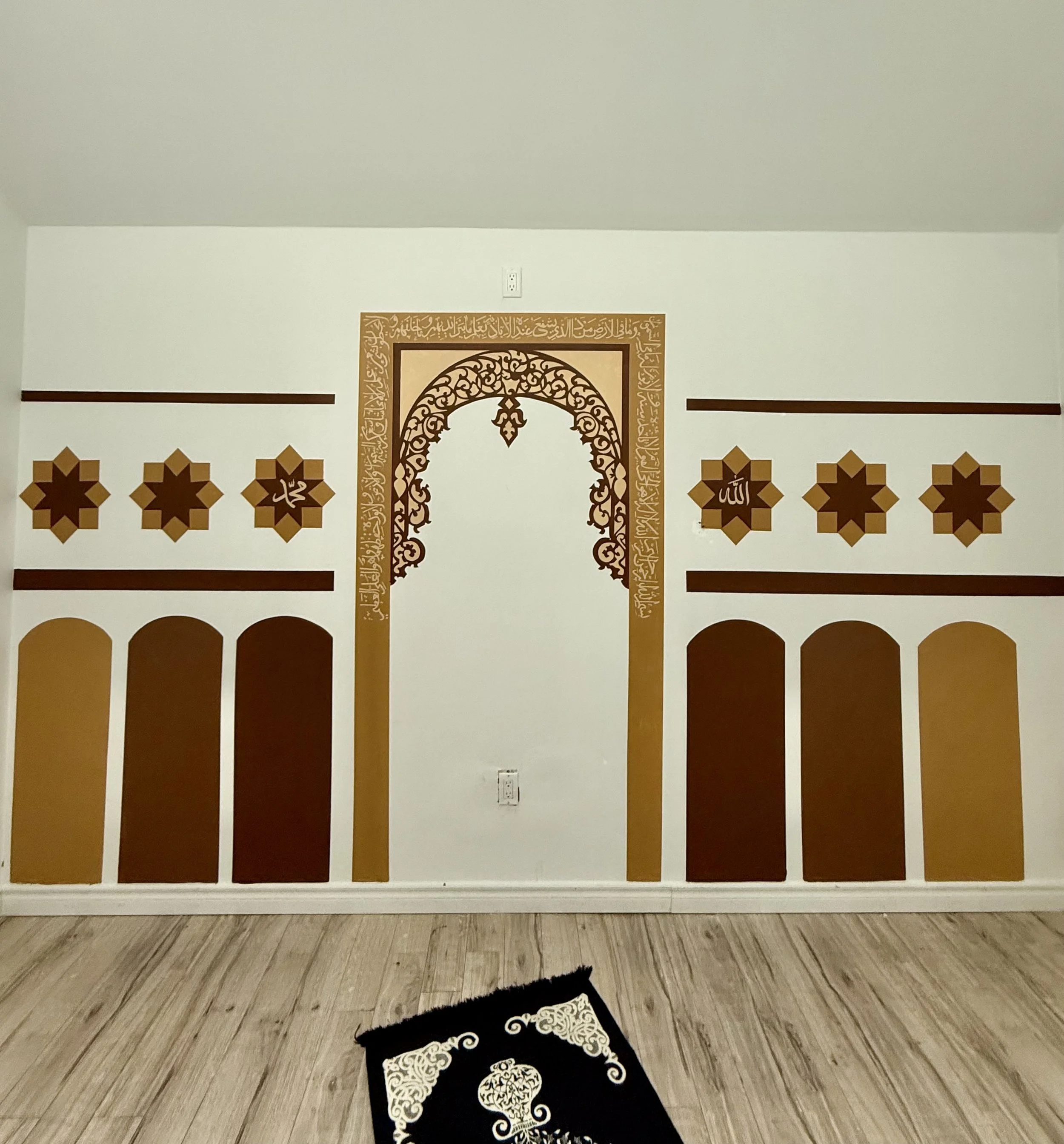 Wall art depicting Islamic architecture with arched doorway, geometric patterns, and Arabic calligraphy in gold and brown on a white wall.