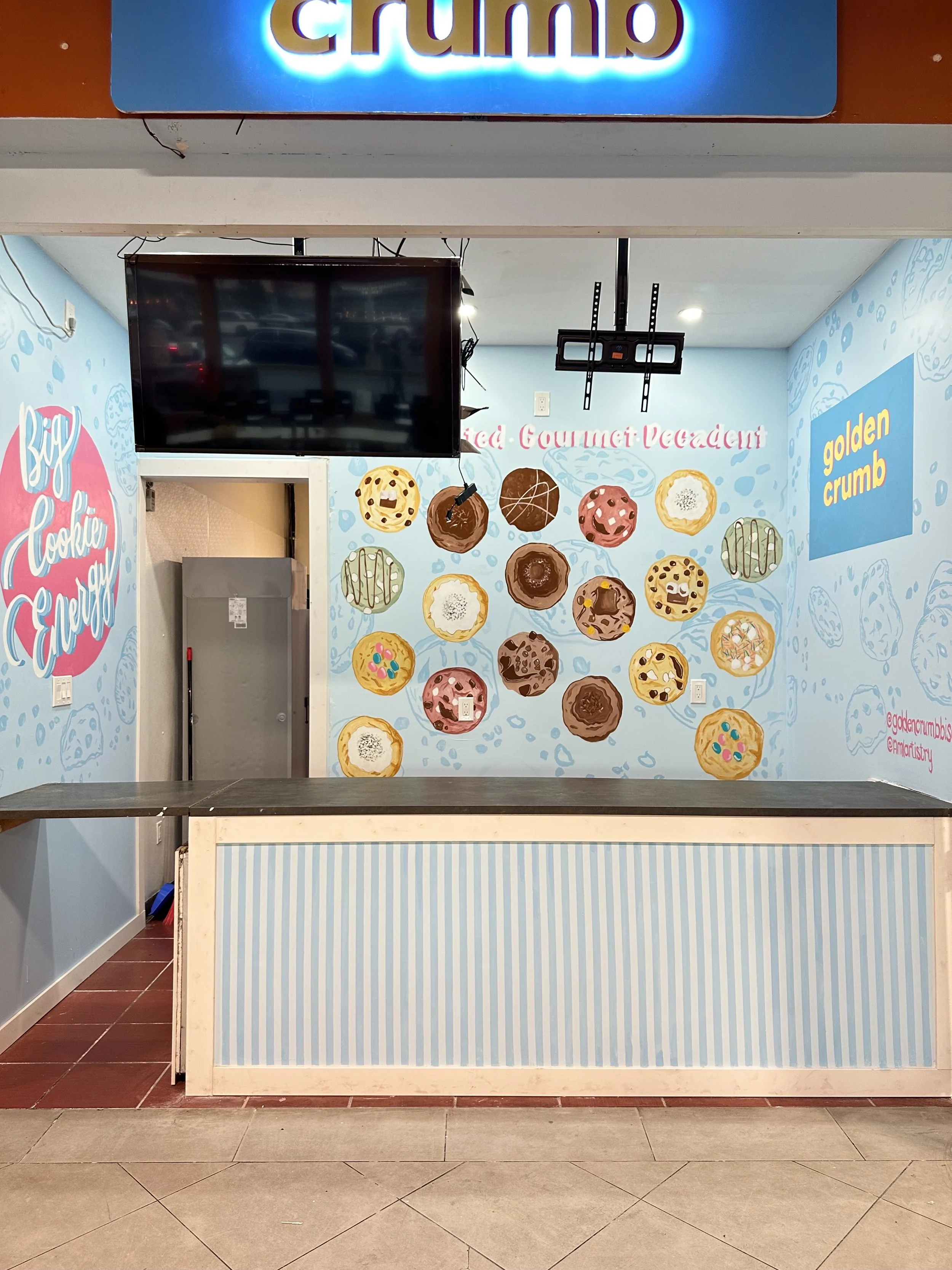 Indoor ice cream shop with a blue and white striped counter, wall mural of various ice cream and cookie illustrations, and a TV mounted on the wall. Sign above the shop says "Grumo."