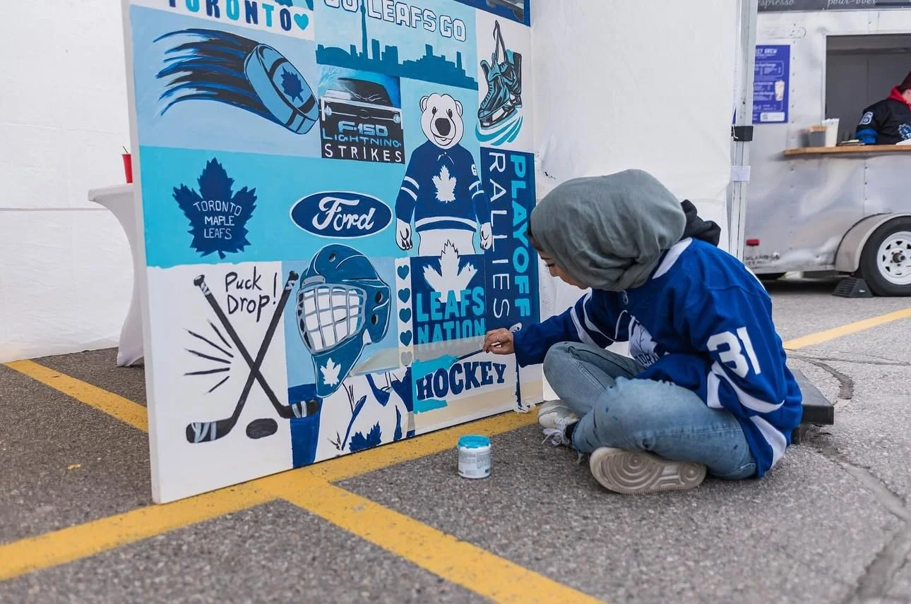 A person wearing a blue hockey jersey and jeans, with a gray hooded sweatshirt, is sitting on the ground and painting a mural featuring Toronto Maple Leafs hockey team symbols and logos. The mural includes images of a hockey puck, face mask, hockey s