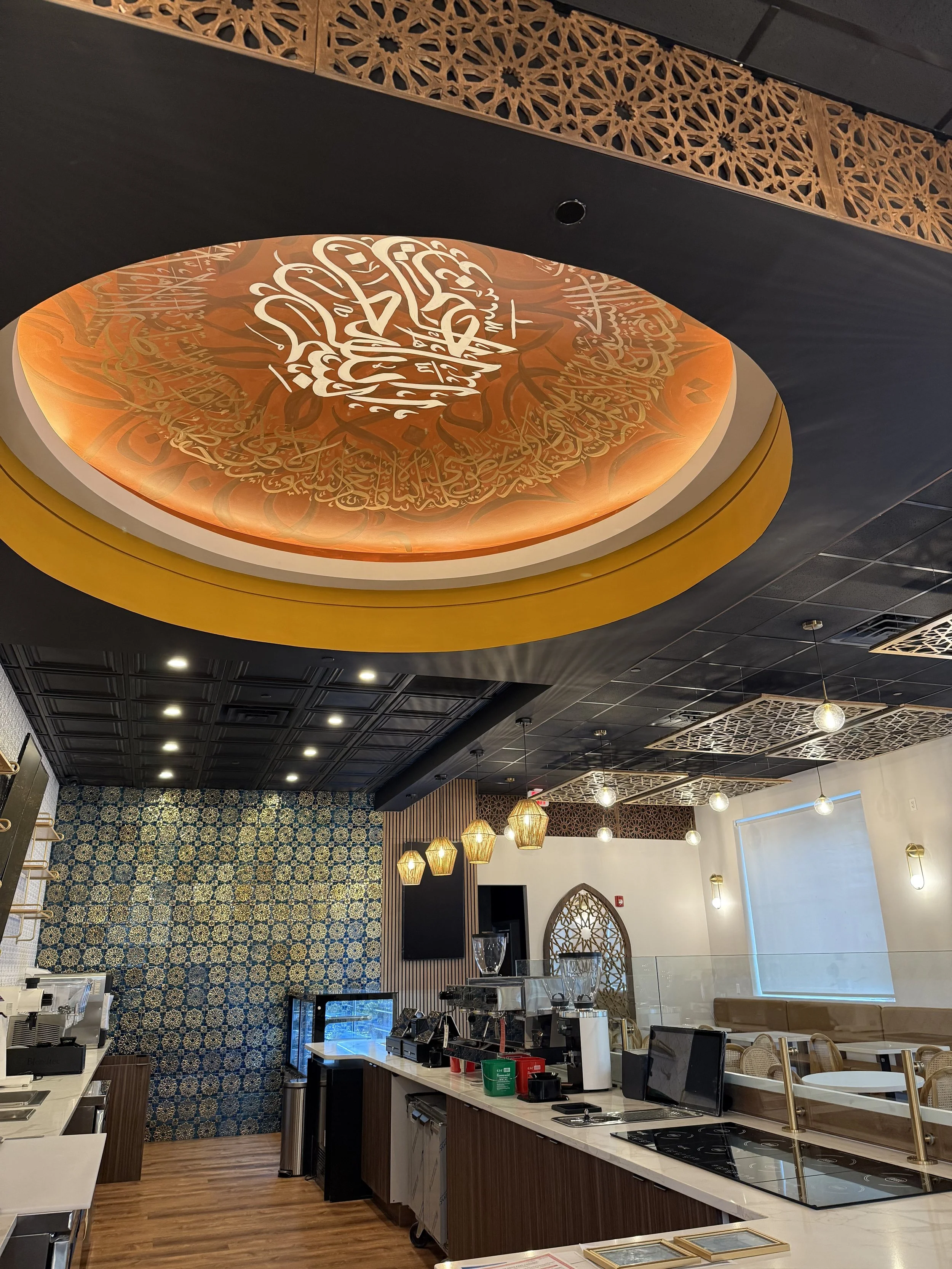 Interior of a restaurant or cafe with a decorative ceiling. The ceiling features a large circular orange and gold piece of Islamic calligraphy. The space has hanging pendant lights, a patterned blue and gold wall, and a counter with coffee machines a