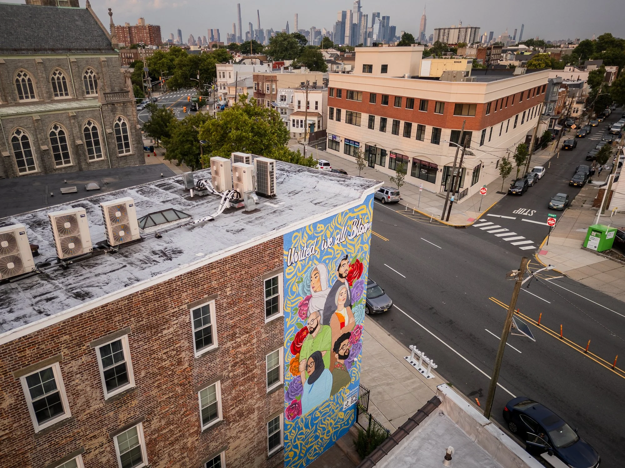 View of a city street with a large mural on a brick building, featuring diverse people and the words 'United We All Belong', with a city skyline in the background.