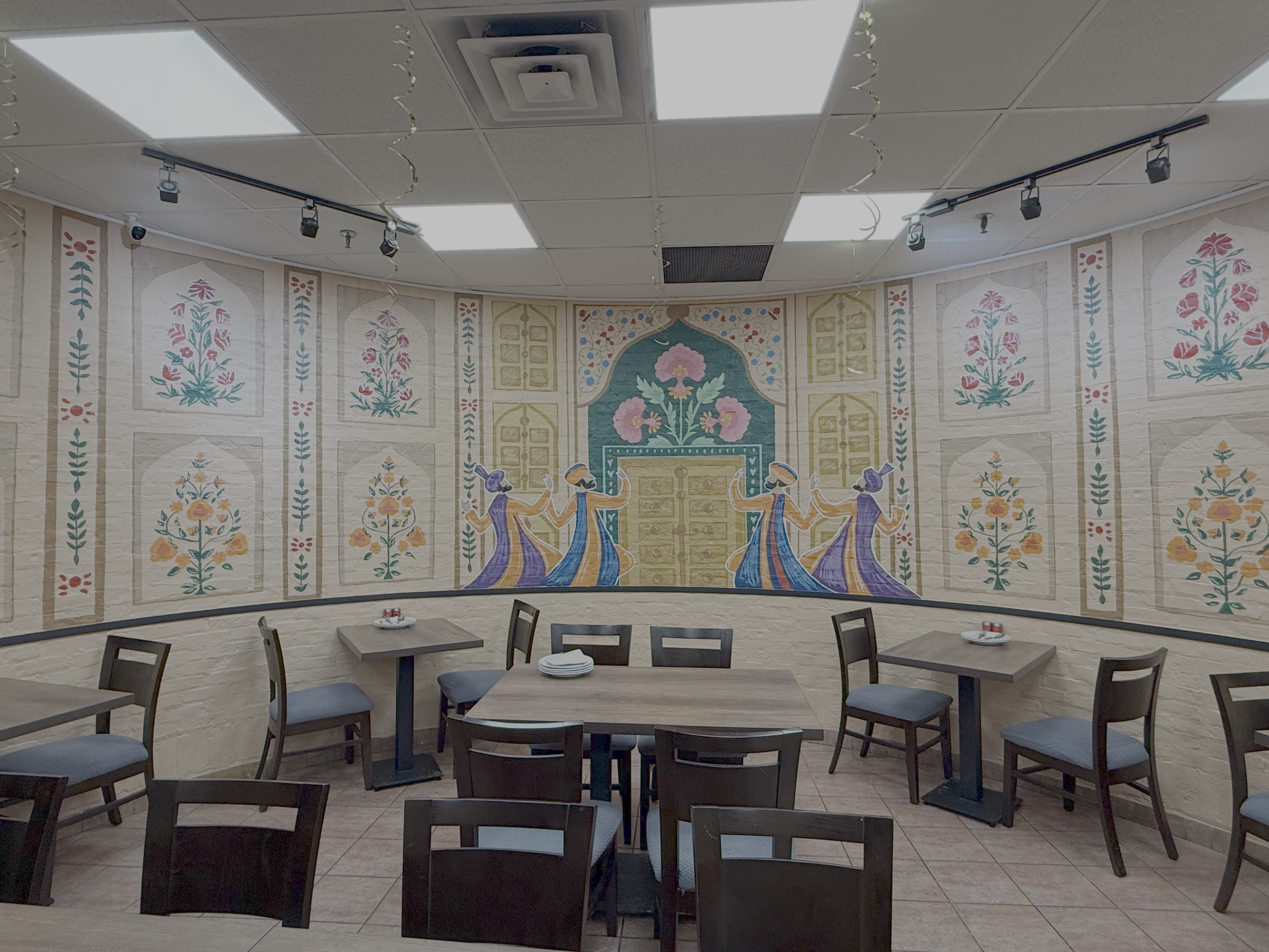 Interior of a restaurant with a decorative mural on the curved wall featuring traditional dancers and floral patterns, wooden tables and chairs, and ceiling lights.