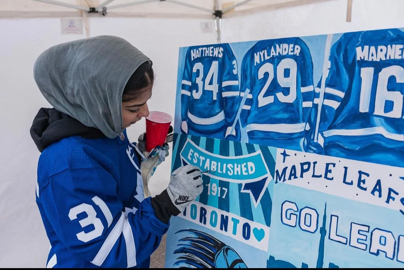 A person in a blue hockey jersey, with a grey hood over their head, is drawing or painting on a large blue hockey-themed mural. The mural features hockey players with numbers and a banner with the text "Established 1917" and "Toronto".