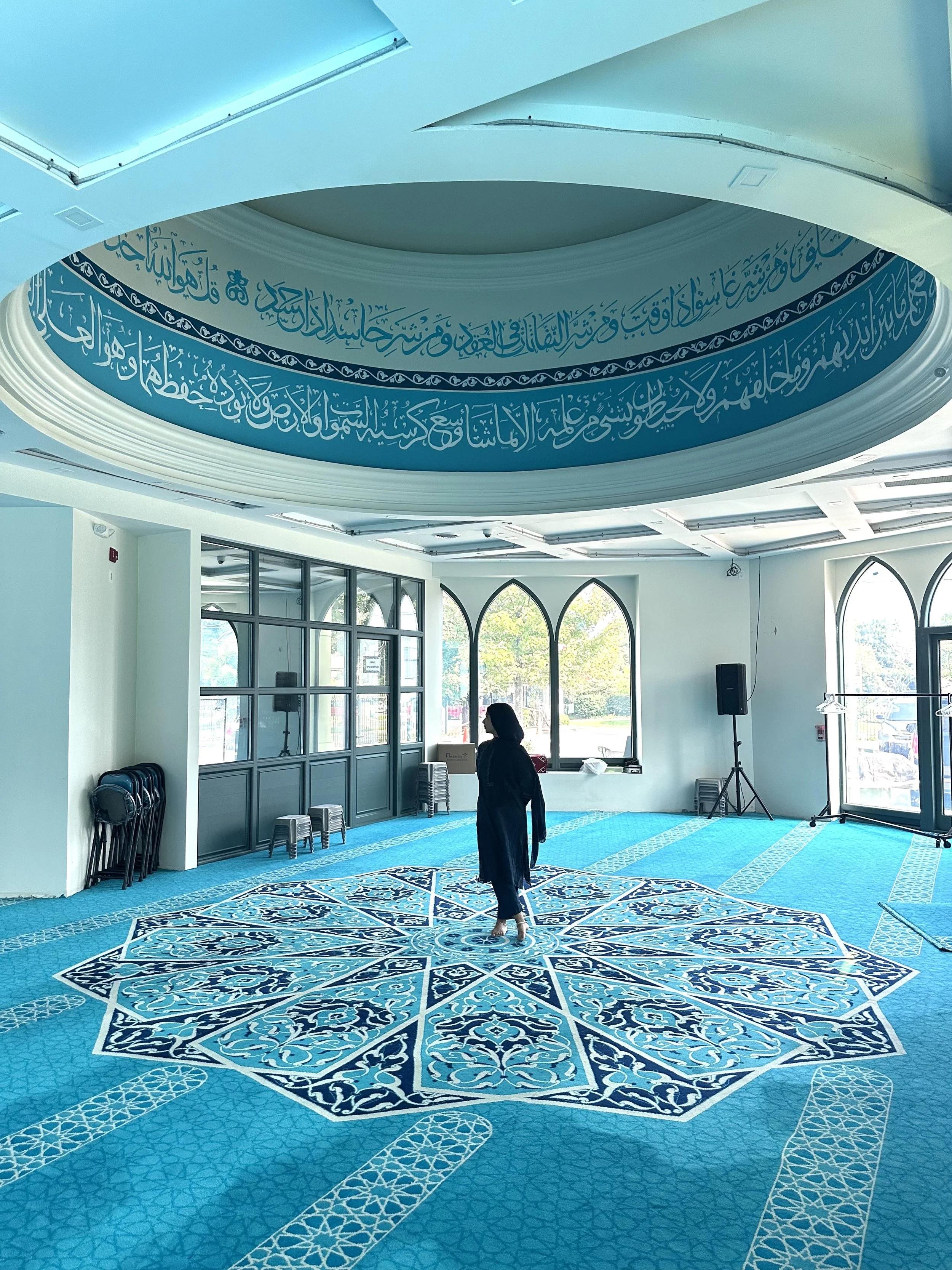 A woman standing alone in a mosque prayer hall with a blue and white patterned carpet, arched windows, and a large circular blue calligraphic decoration on the ceiling.