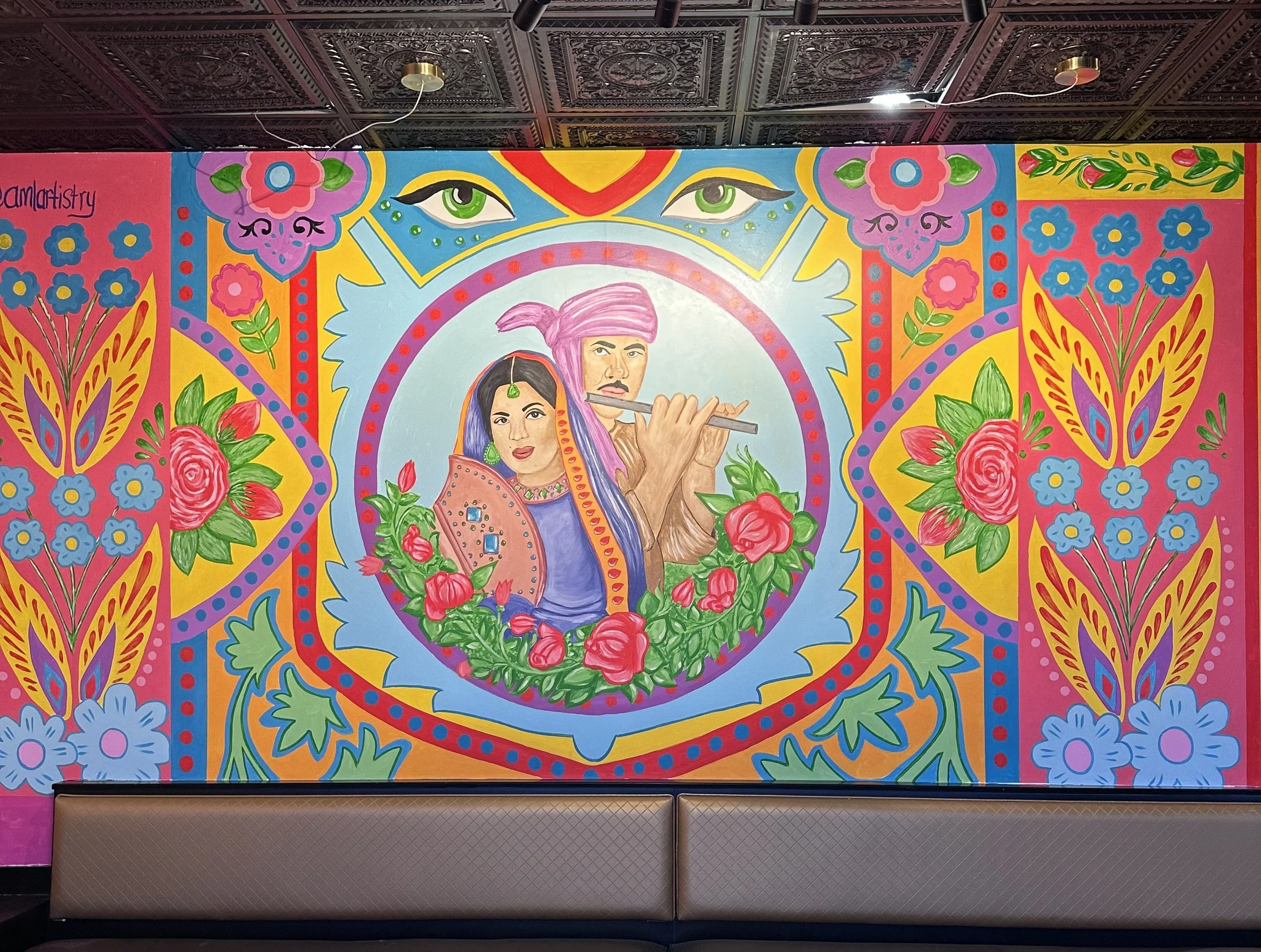 Colorful mural depicting a man and woman dressed in traditional Indian attire, surrounded by flowers and decorative patterns, with a pair of stylized eyes at the top.