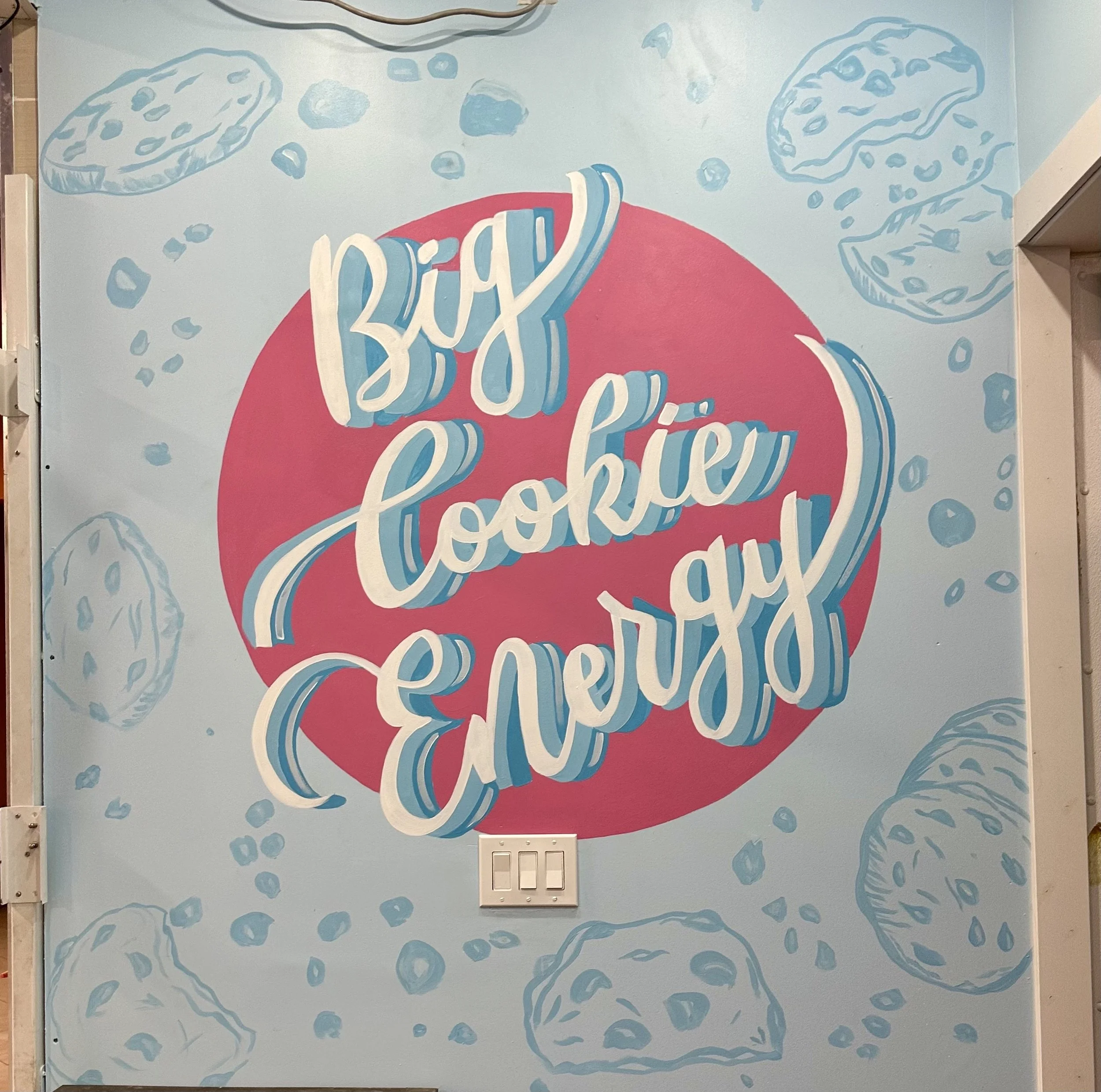 Decorative wall with the phrase 'Big Cookie Energy' in colorful, stylized lettering surrounded by illustrations of cookies and cookie crumbs.