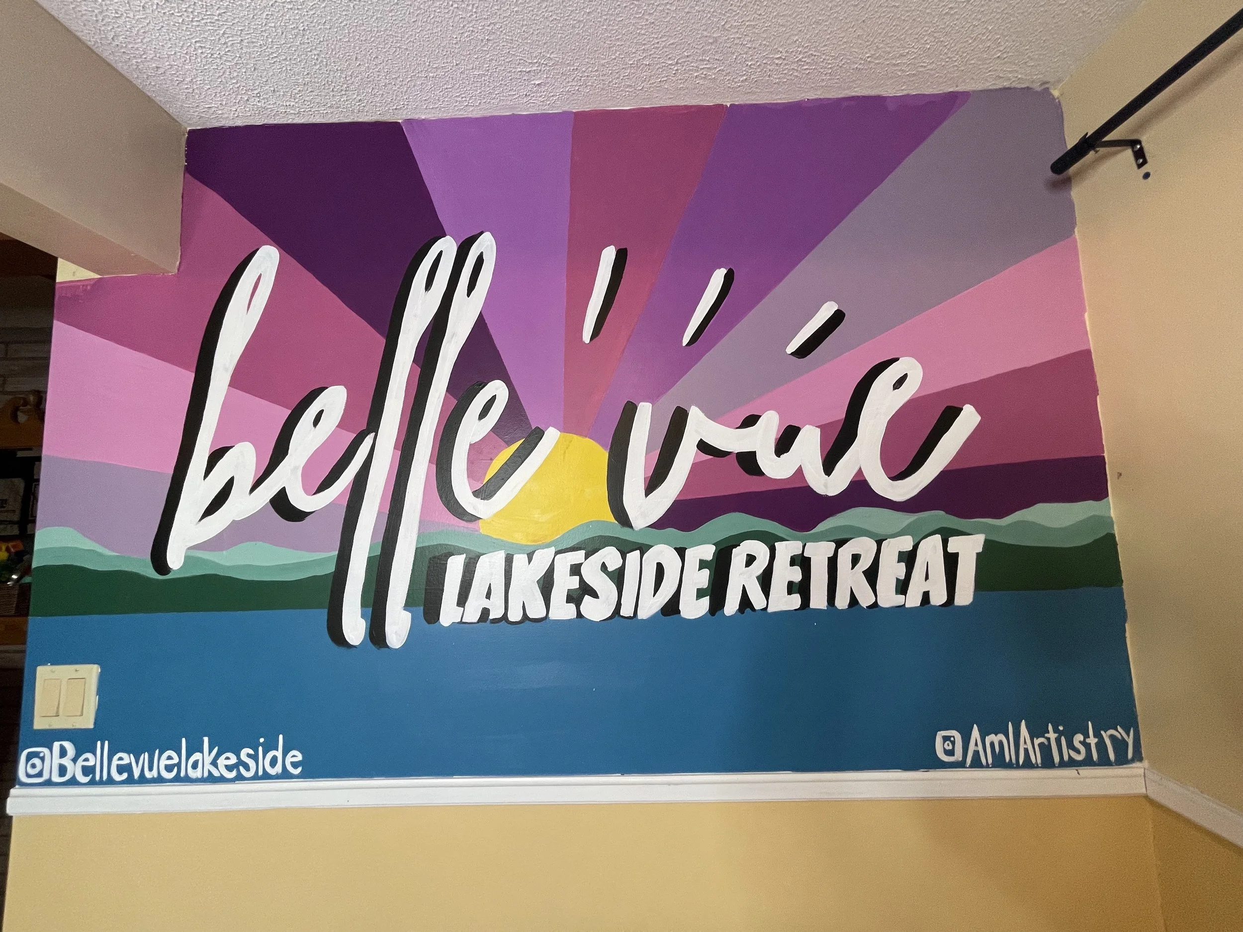 Colorful wall mural with a sunset over lakes and mountains, reading 'hello val! LAKESIDE RETREAT,' with social media handles '@Bellevuelakeside' and '@AmlArtistry'.