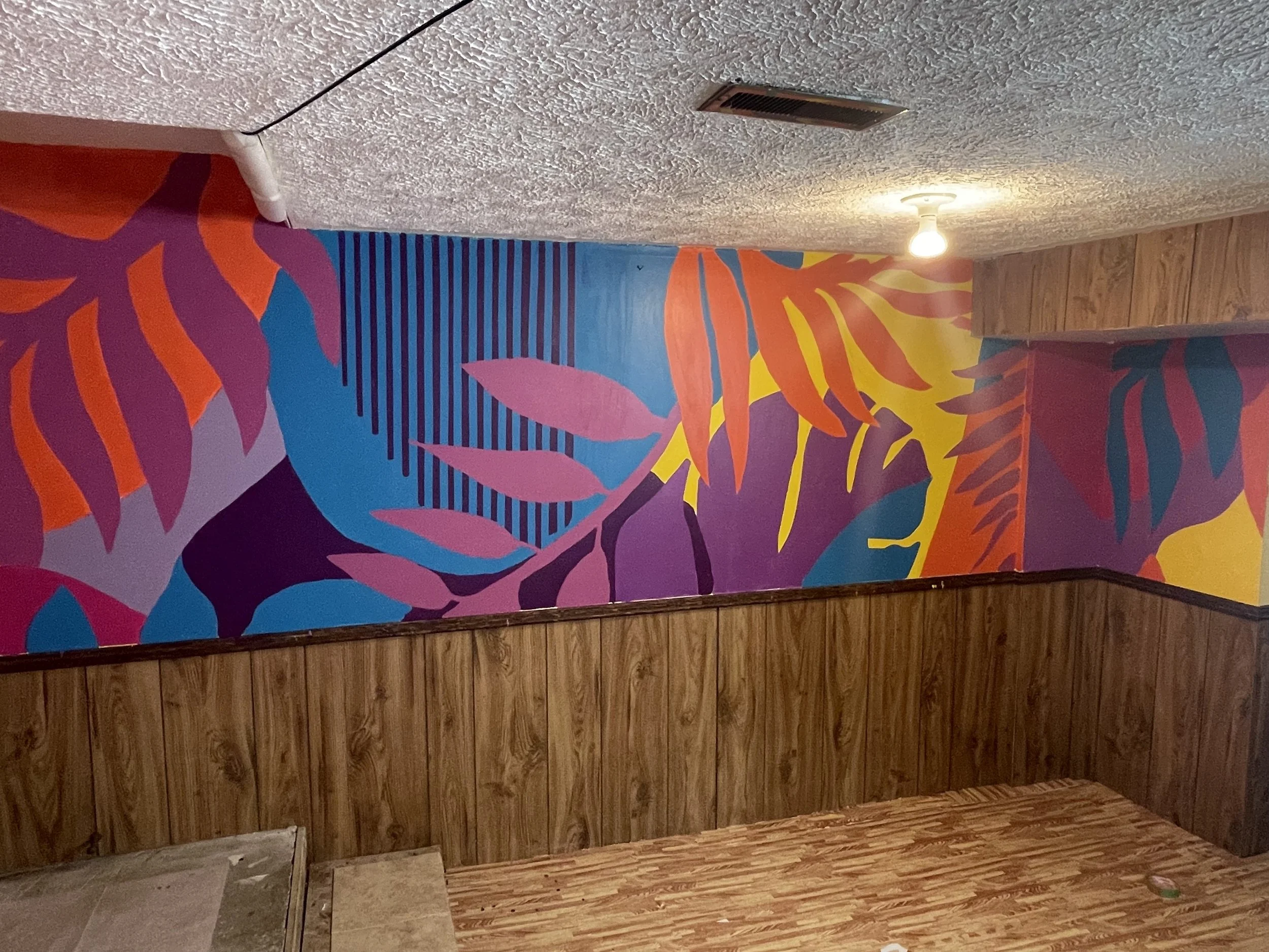Colorful mural with abstract tropical leaves and plant shapes painted on a wall in a room with wooden paneling.
