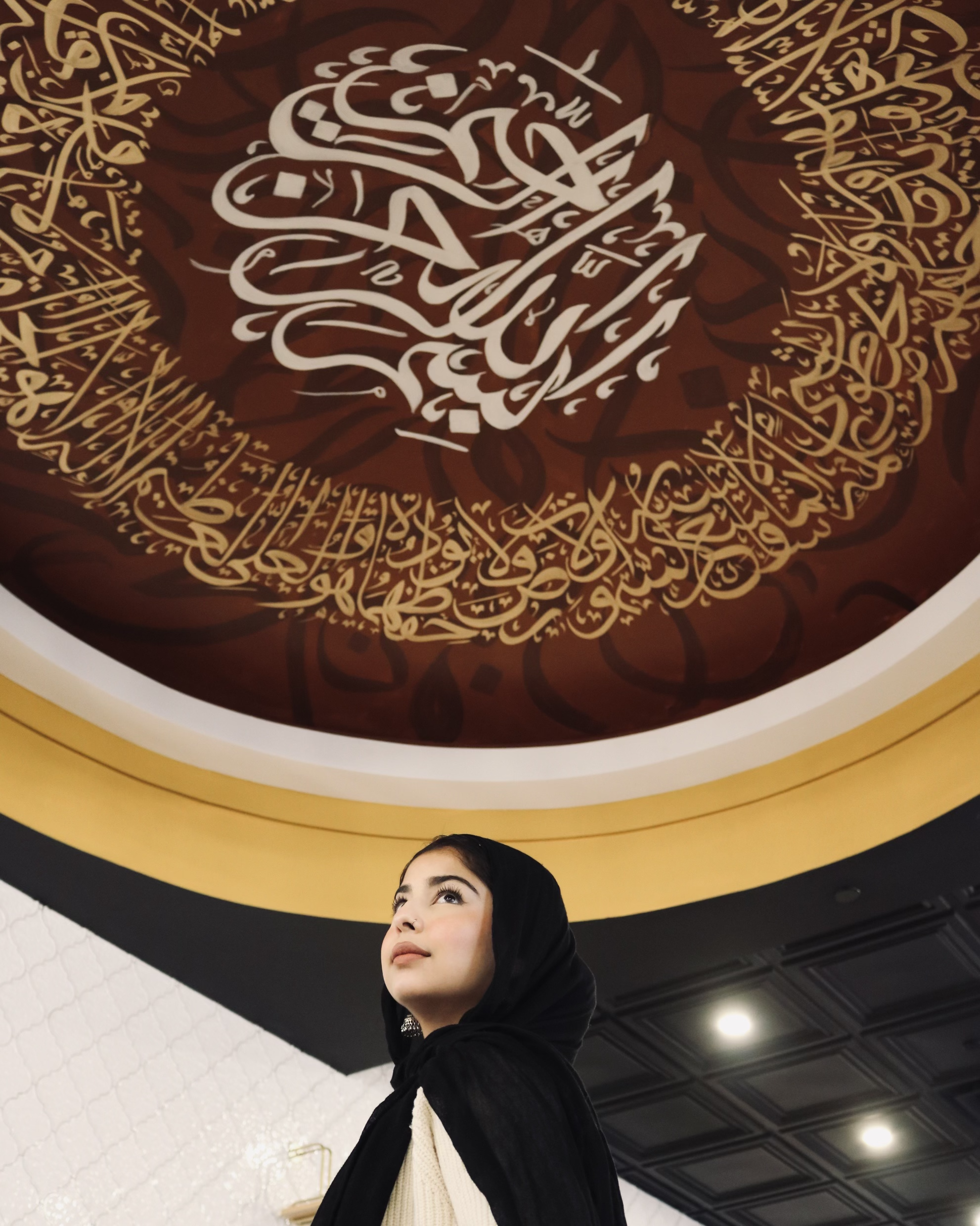 A young woman wearing a black headscarf looking up at a large Islamic calligraphy artwork on the ceiling featuring Arabic script in gold and white, with a dark red background.