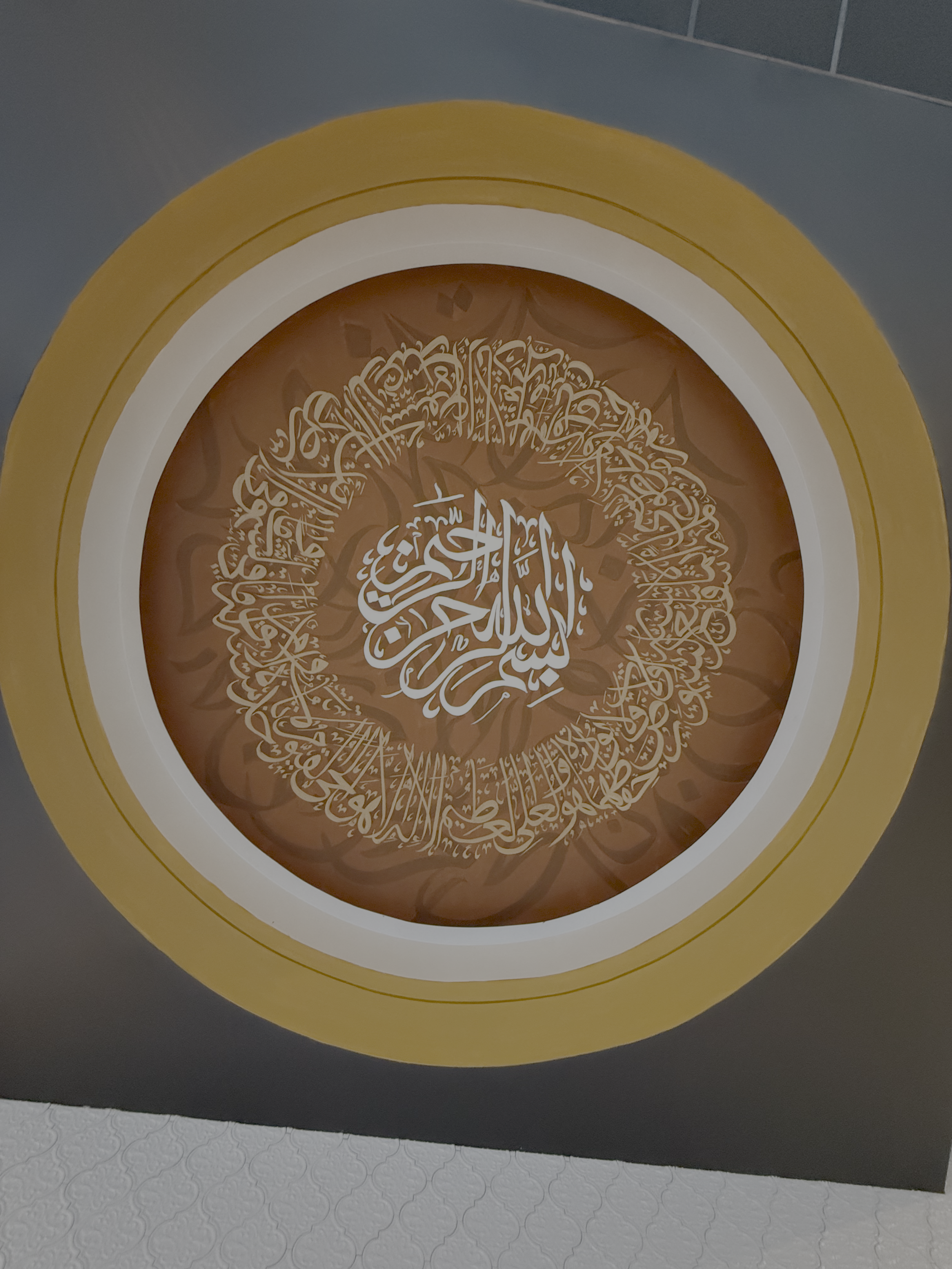 Islamic calligraphy artwork in a circular design, featuring Arabic script, with a beige and brown color scheme on a wall.