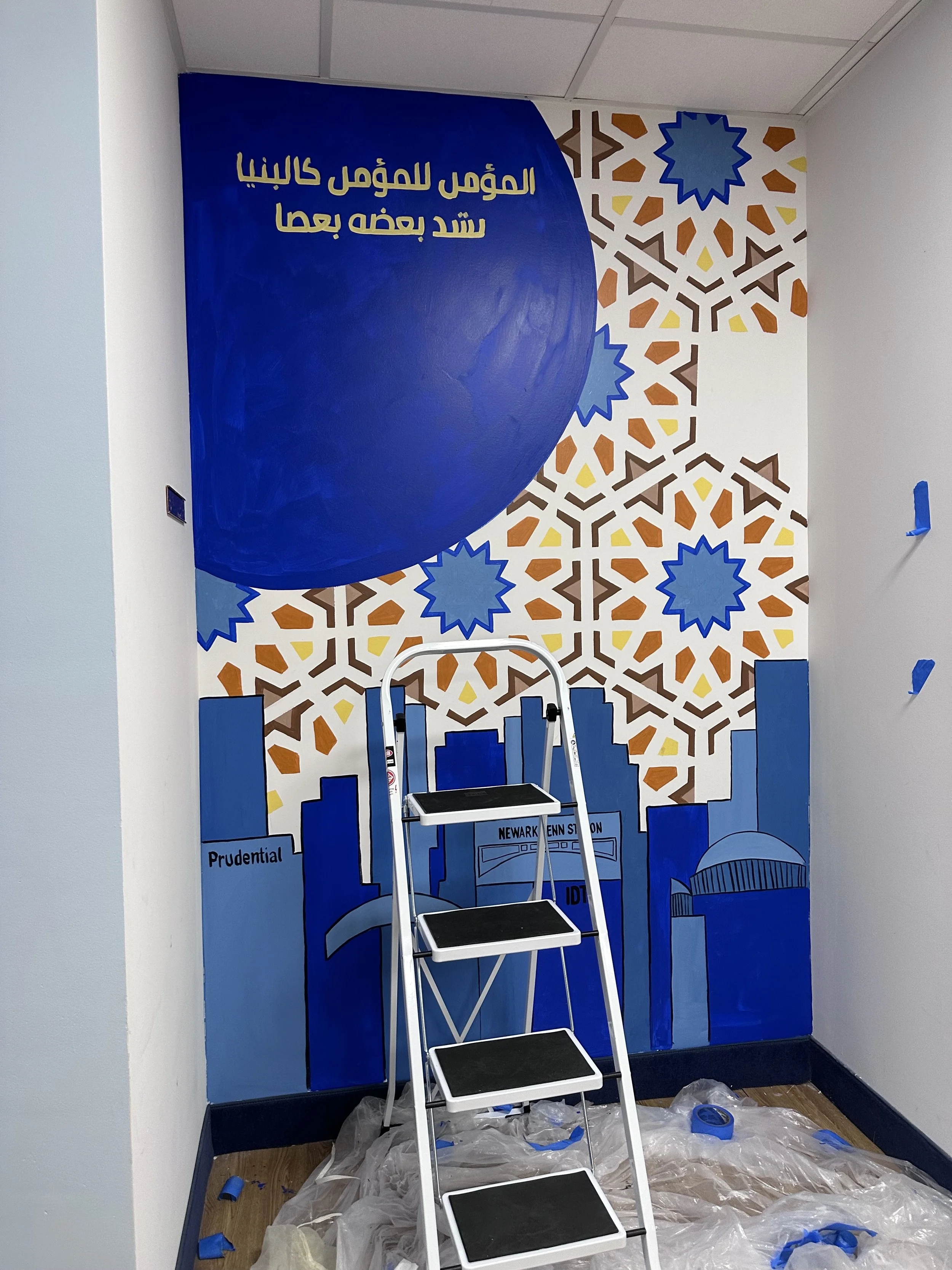 A mural in progress featuring a city skyline, blue geometric designs, and Arabic text, with a step ladder and painter's plastic on the floor.